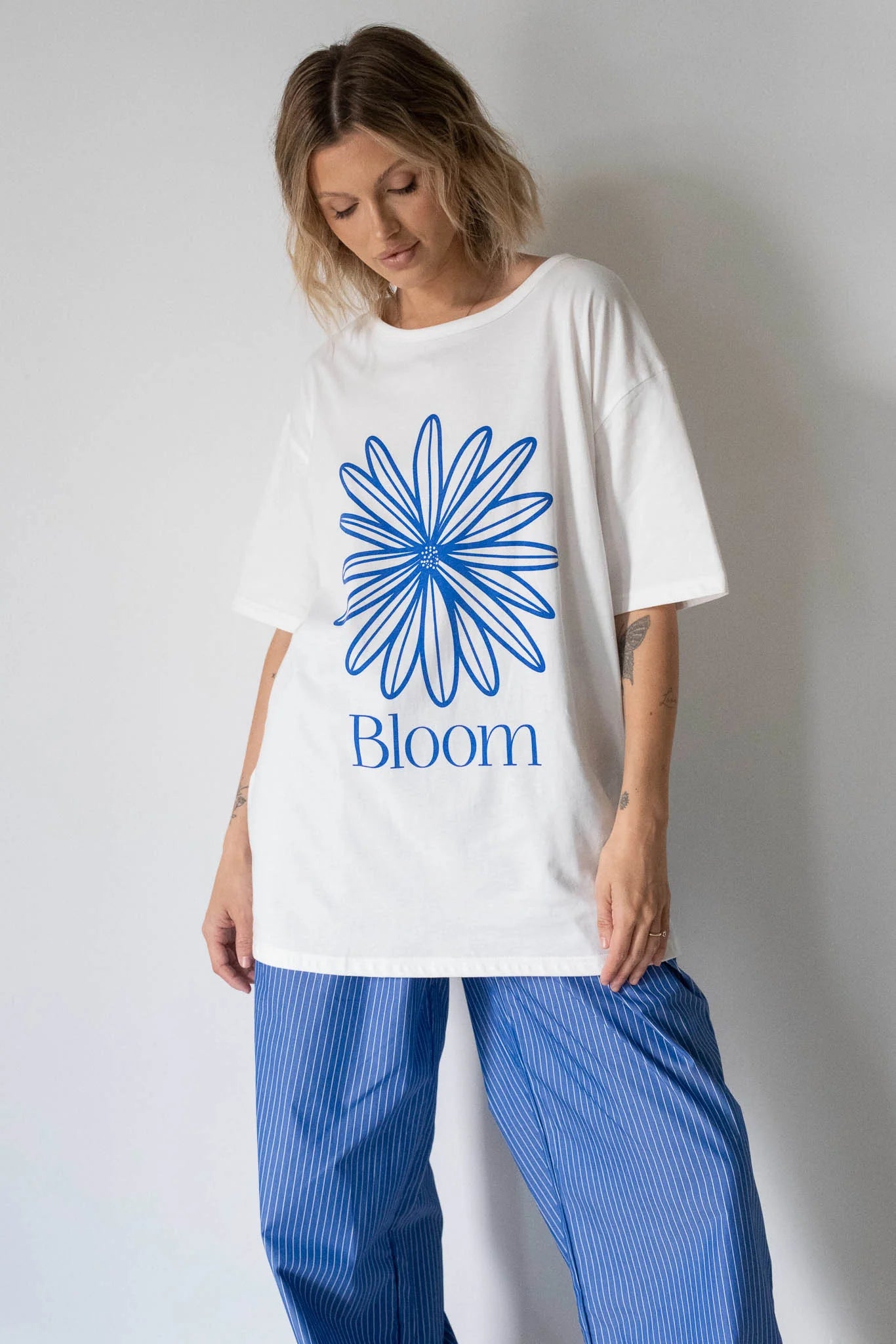A woman wears the Tiny Trove - Women's Bloom Relaxed Tee in white/blue by TINY TROVE, featuring an oversized fit with a large blue abstract flower and "Bloom" logo on the front, paired with blue striped pants against a light background.
