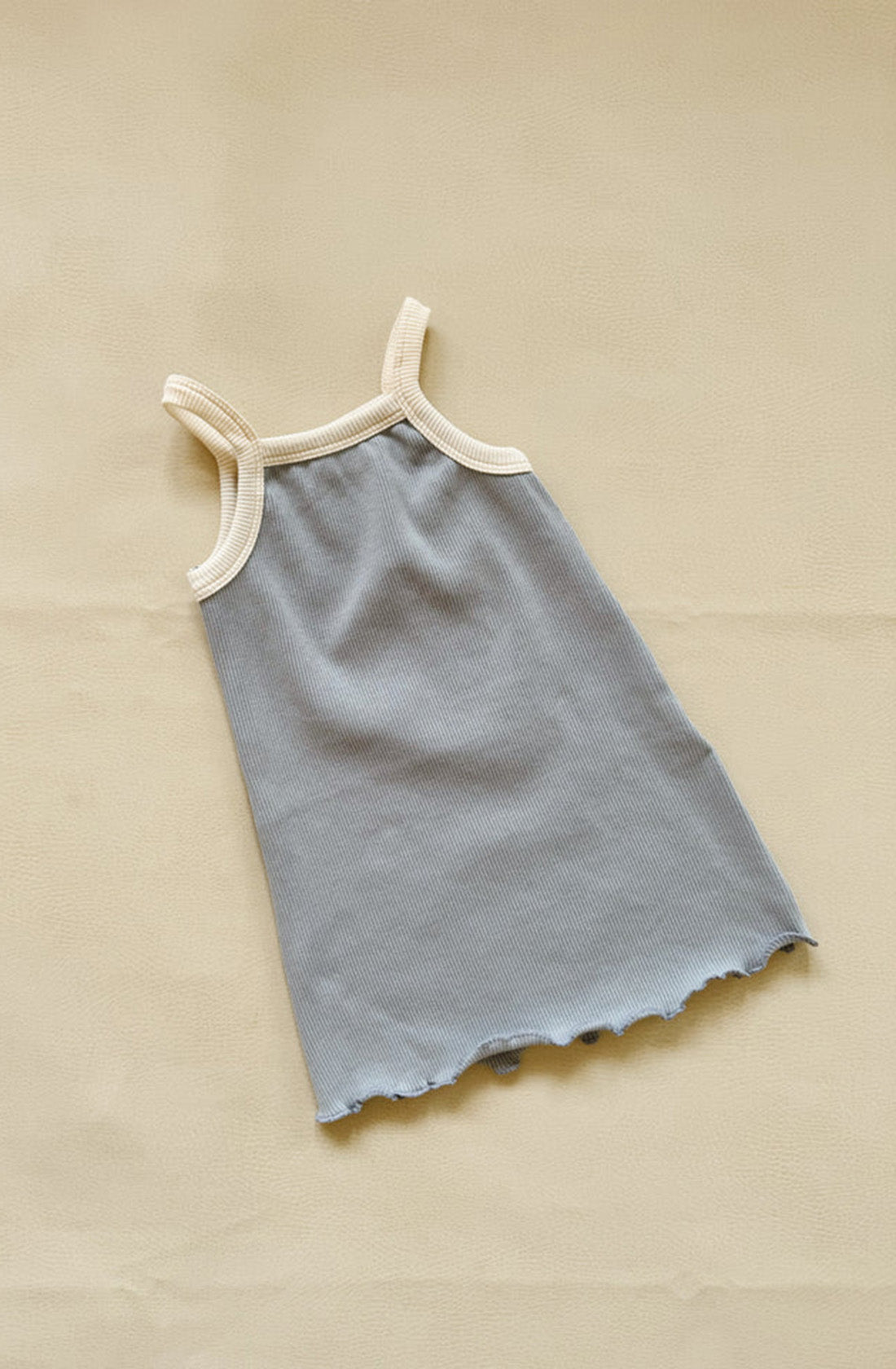 The Elsie Dress by TINY TROVE, primarily blue and cream with a sleeveless ribbed cotton design, features beige trim around the neck and armholes. It also showcases a ruffle detail at the hem and is laid out flat on a beige surface.