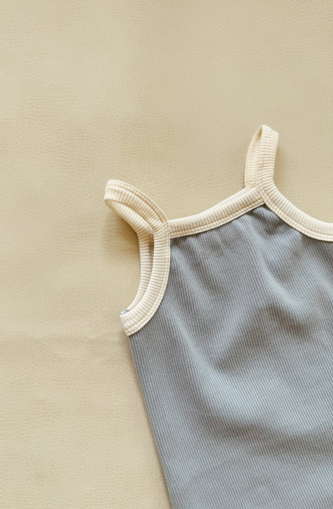 A minimalist image of the Elsie Dress Ivory Blue/Cream by TINY TROVE in muted gray with white trim, laying flat on a beige textured surface. Only the upper half of the dress is visible, featuring its shoulder straps and neckline with subtle ruffle detail.