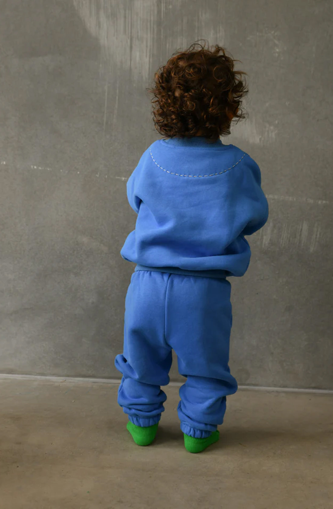 A child with curly hair is standing with their back to the camera, dressed in a matching TINY TROVE Woodie 3D Logo Tracksuit in Ocean blue, along with green socks. The comfortable wear contrasts subtly against the plain, textured gray wall in the background.