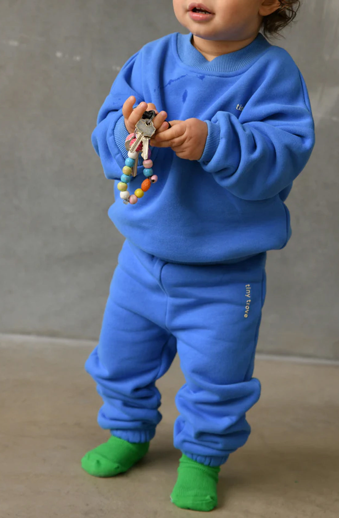 A toddler dressed in the Woodie 3D Logo Tracksuit Ocean by TINY TROVE holds a small beaded toy and a doll while standing indoors. Their comfortable outfit is paired with green socks, set against a plain grey wall.