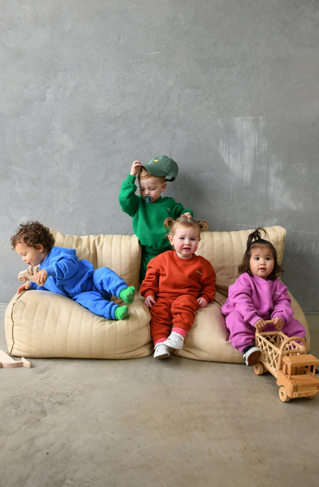 Four toddlers sit on a beige cushion against a gray wall, each dressed in colorful, comfortable wear: blue, green, red, and purple fleece cotton outfits. The toddler in green is adjusting a hat from the TINY TROVE Woodie 3D Logo Tracksuit Ocean collection, while the one in purple holds a wooden toy stroller. They all have different playful expressions.