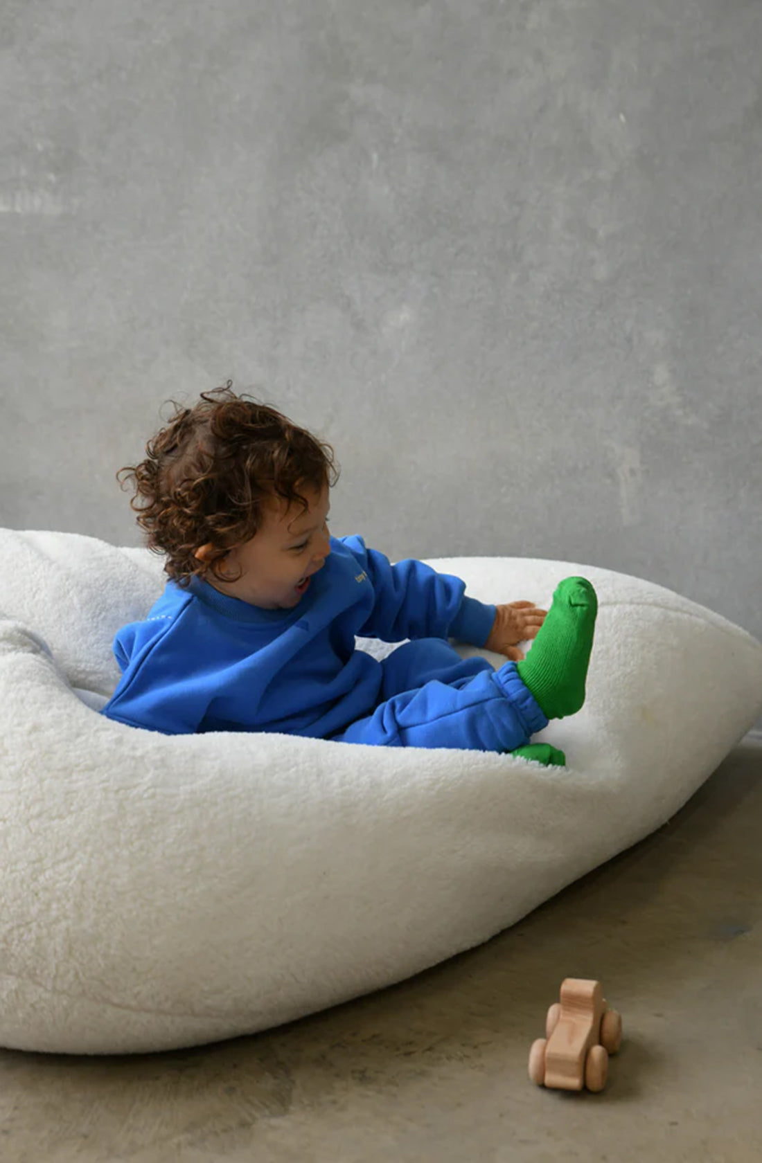 A young child with curly brown hair, wearing the Woodie 3D Logo Tracksuit Ocean by TINY TROVE and green socks, sits on a large, white bean bag chair. The child is looking at their green sock with interest. A small wooden toy is visible on the floor nearby. The background is a plain, light gray wall.