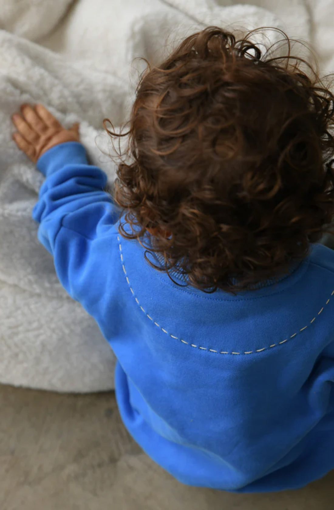 A toddler with curly hair wears the TINY TROVE Woodie 3D Logo Tracksuit Ocean while touching a soft, fluffy white blanket. The child is turned away, with the back of their head and hand visible. The scene appears to be indoors, highlighting the comfortable wear and cozy feel of fleece cotton surroundings.