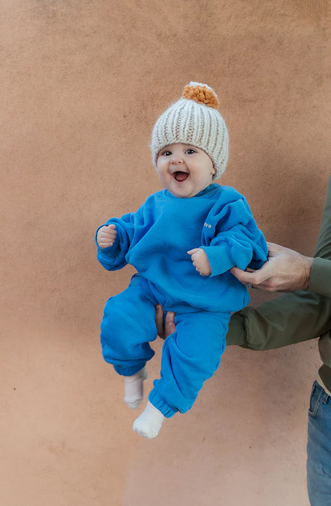 A cheerful baby in a comfortable **Woodie 3D Logo Tracksuit Ocean** by **TINY TROVE** and a white knit hat with an orange pom-pom is being held up against a peach-colored background. The baby, wearing cozy fleece cotton, has a joyful expression with a wide smile and outstretched arms. Part of an adult’s arm and hand are visible.