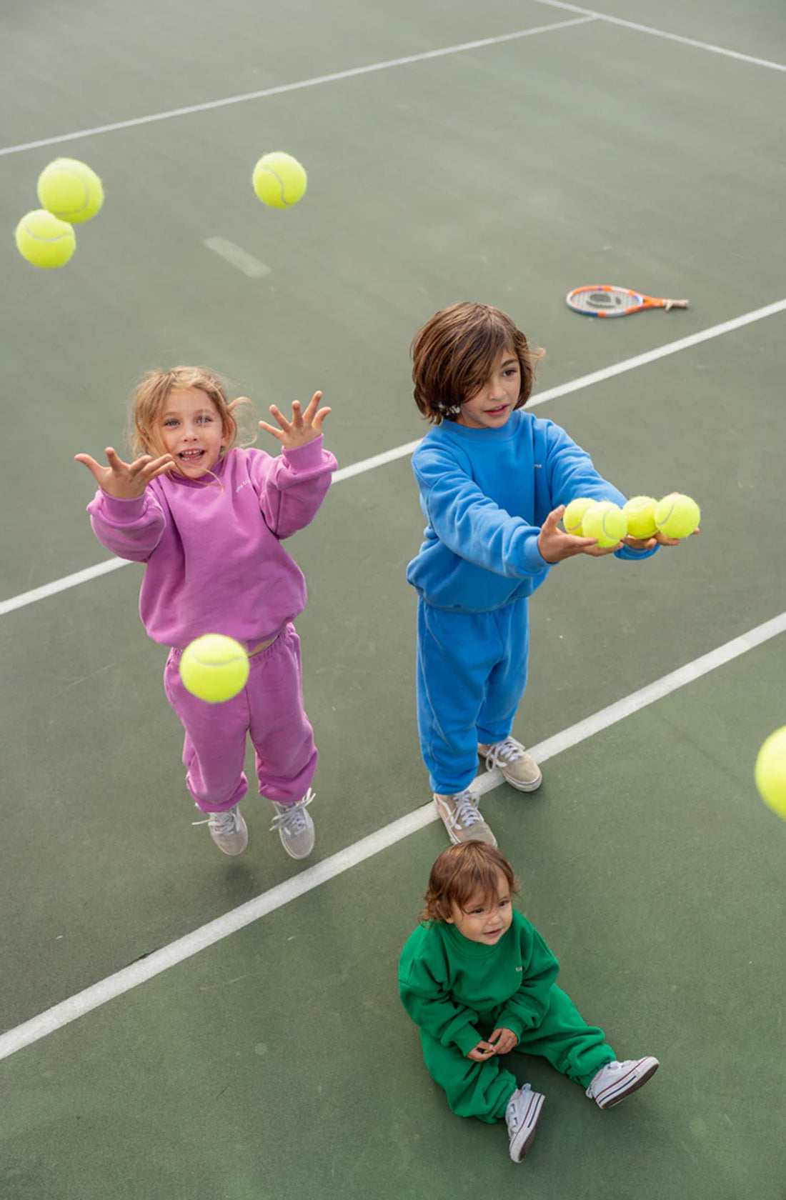 Here's the rewritten sentence using the given product data:

Three children are playing on a tennis court. Two older children, dressed in TINY TROVE Woodie 3D Logo Tracksuits — one in pink and one in blue adorned with 3D logos — are joyfully throwing multiple tennis balls into the air. A younger child in a green fleece cotton tracksuit sits on the ground holding a tennis ball, with a racket lying nearby.