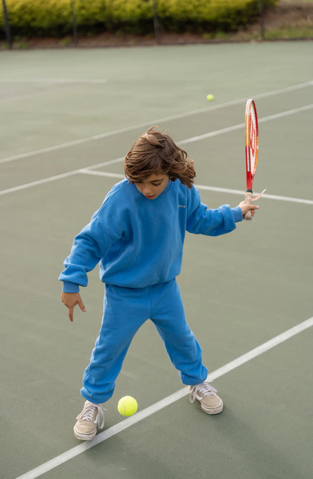 A young child wearing the Woodie 3D Logo Tracksuit Ocean from TINY TROVE and beige sneakers is playing tennis on an outdoor court. The child is holding a tennis racket and focused on hitting a ball on the ground, with greenery seen in the background.