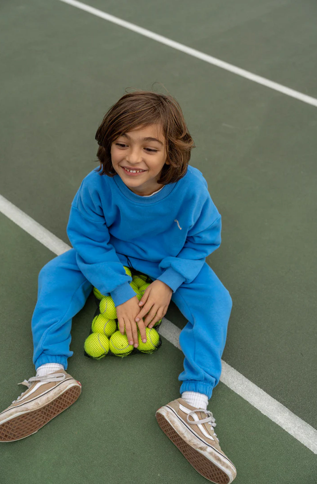 A young child with shoulder-length brown hair, wearing a Woodie 3D Logo Tracksuit Ocean by TINY TROVE and white sneakers, sits on a tennis court. The child is smiling and holding multiple yellow tennis balls between their legs. The court's green surface and white lines are visible.