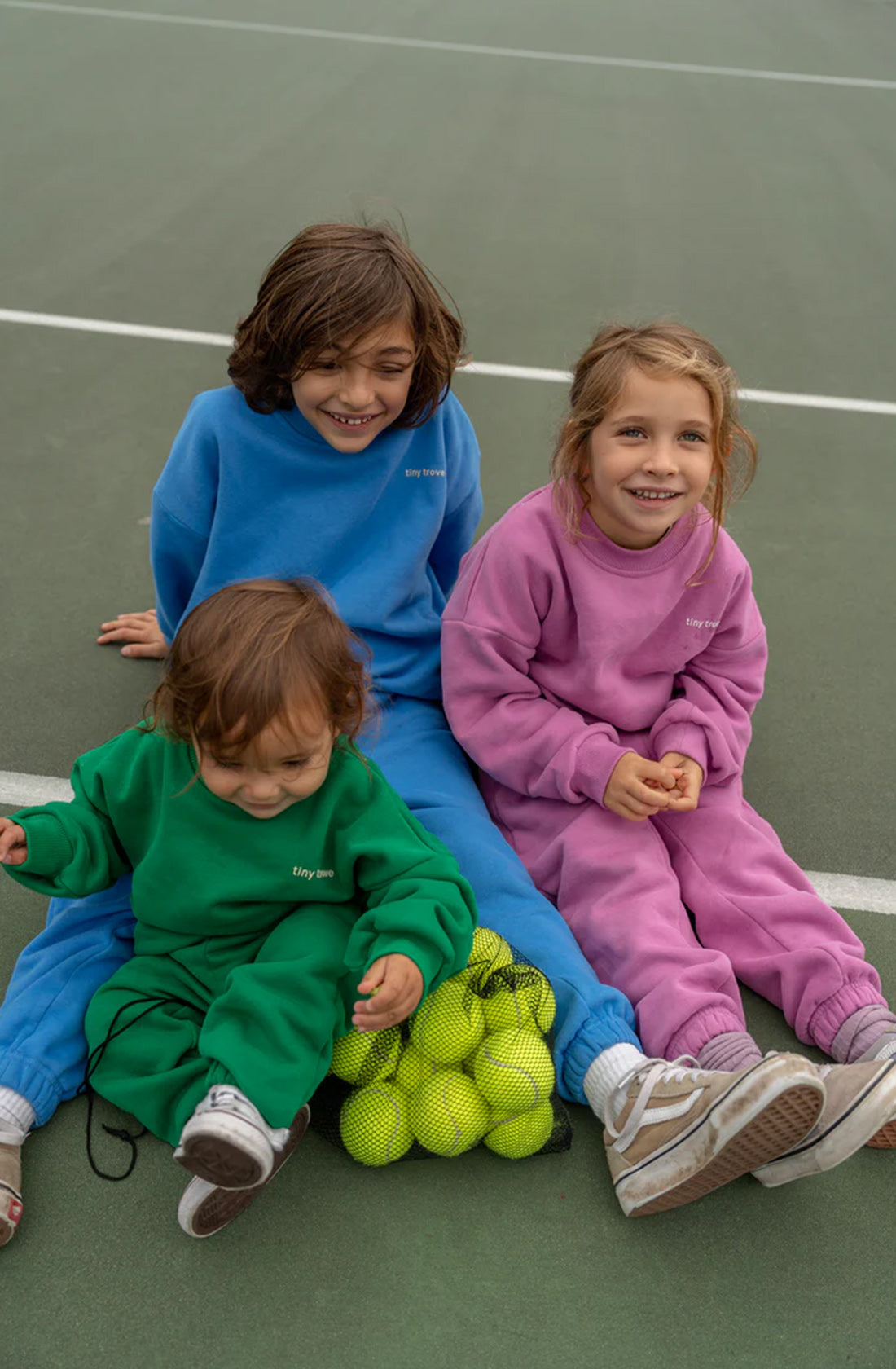 Three children dressed in colorful tracksuits and comfortable wear sit on a tennis court. The child on the left is wearing a green tracksuit, the center child sports the TINY TROVE Woodie 3D Logo Tracksuit Ocean, and the one on the right is in a pink tracksuit. They are sitting next to a pile of tennis balls.