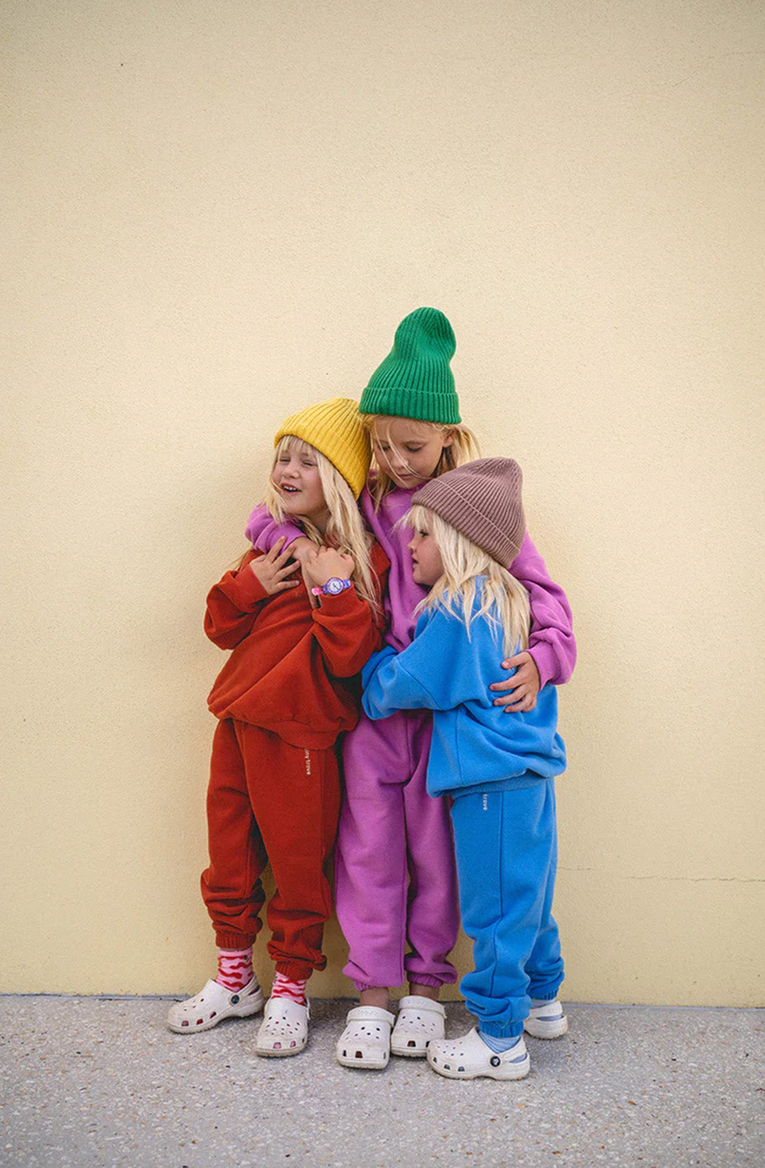 Three children stand in a group hug against a pale yellow wall. Each child wears a colorful Woodie 3D Logo Tracksuit from TINY TROVE along with a beanie: one in red, one in pink, and one in blue ocean color. They all have light-colored hair and are wearing Crocs on their feet. The tracksuits are made of cozy fleece cotton, ideal for comfortable wear.