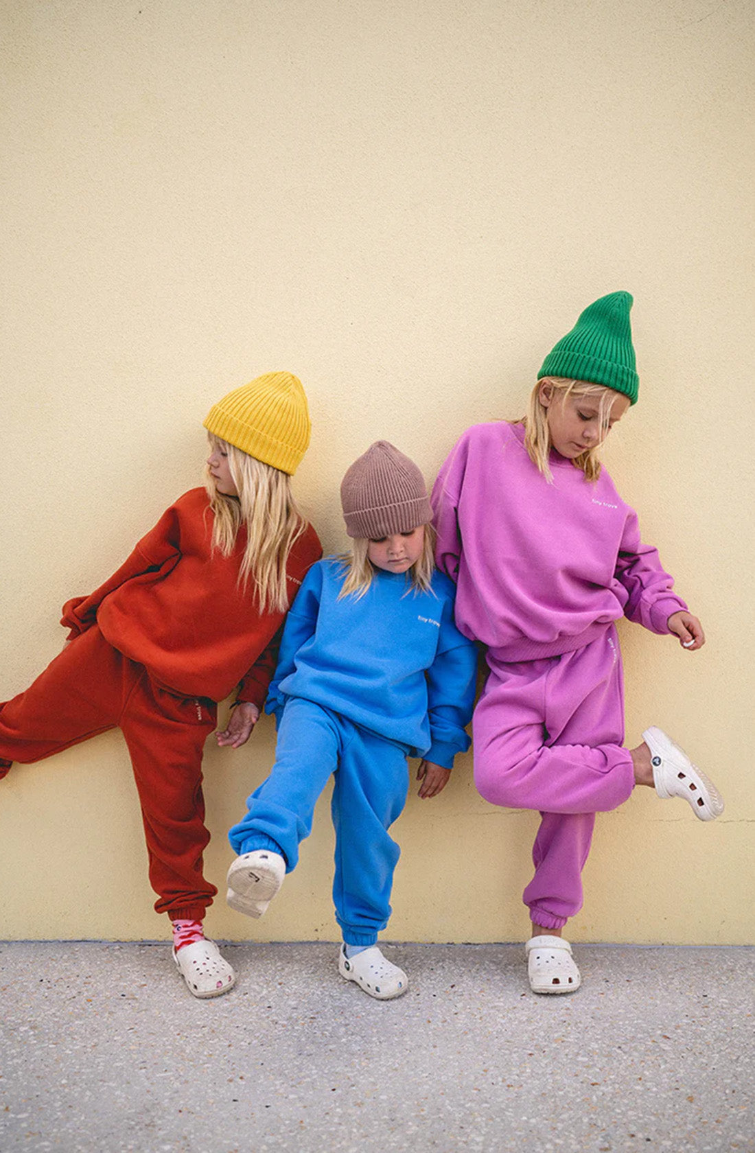 Standing against a plain wall, three children showcase the comfortable Woodie 3D Logo Tracksuit Ocean by TINY TROVE. The child on the left wears a vibrant red tracksuit, the middle child is dressed in serene blue, and the child on the right sports a soft pink outfit. Each pair their attire with cozy knit beanies and white Crocs featuring eye-catching 3D logos as they playfully pose for the camera.