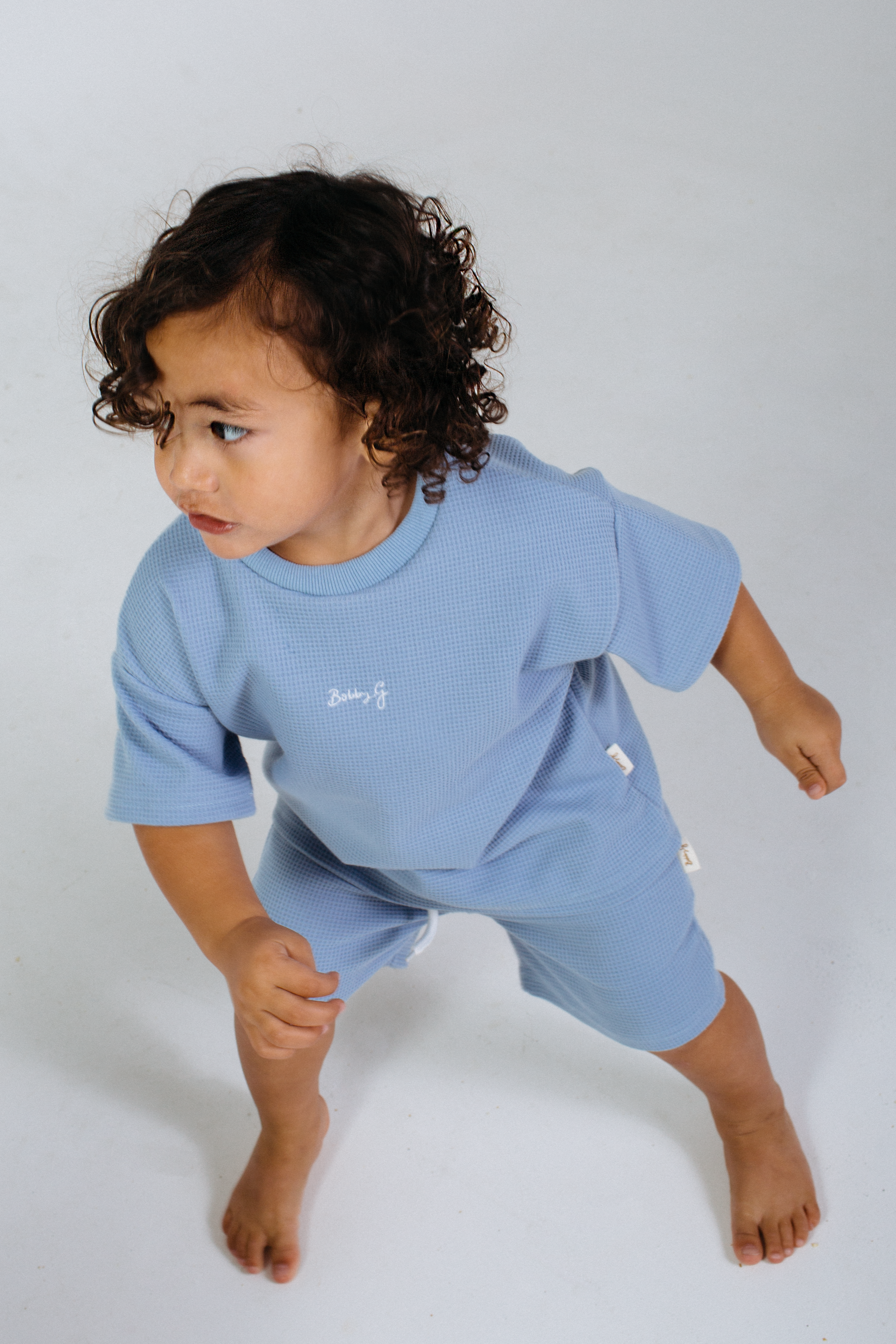 A young child with curly hair is wearing a matching light blue outfit featuring a subtle Bleu texture. The child, possibly dressed in the Ziggy Tee Bleu from BOBBY G BABY WEAR, is barefoot and seems to be moving against a plain white background, gazing off to the side.