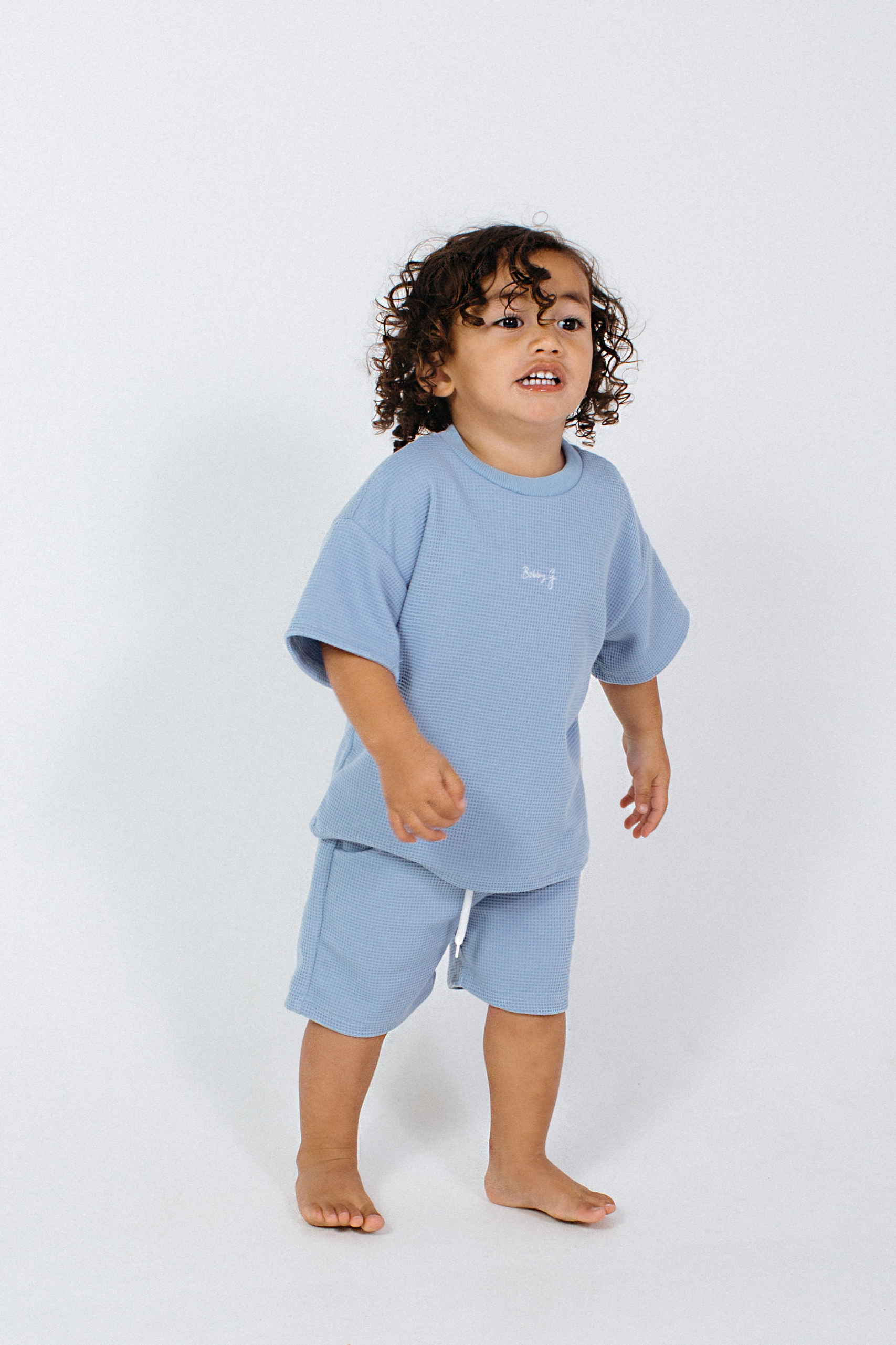 A toddler with curly hair stands barefoot, wearing a light blue short-sleeve top along with Frankie Shorts Bleu from BOBBY G BABY WEAR. The plain white background highlights the child's curious and joyful expression, enhancing the stylish and comfortable vibe conveyed by the luxe waffle fabric.