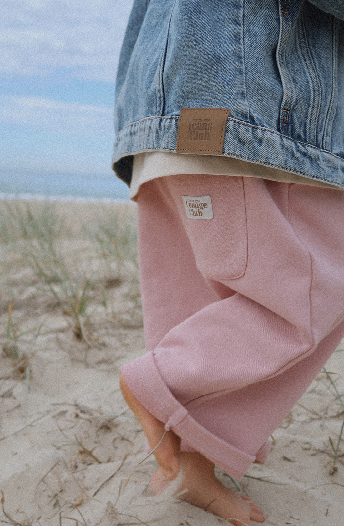 A person with rolled-up TWIN COLLECTIVE Lounge Track Pants in Pink Haze and a light denim jacket walks barefoot on a sandy beach. The denim jacket features a brown patch with "Jeans Club" on it, and the track pants have a "Lounge Club" tag. The scene captures a relaxed coastal atmosphere.