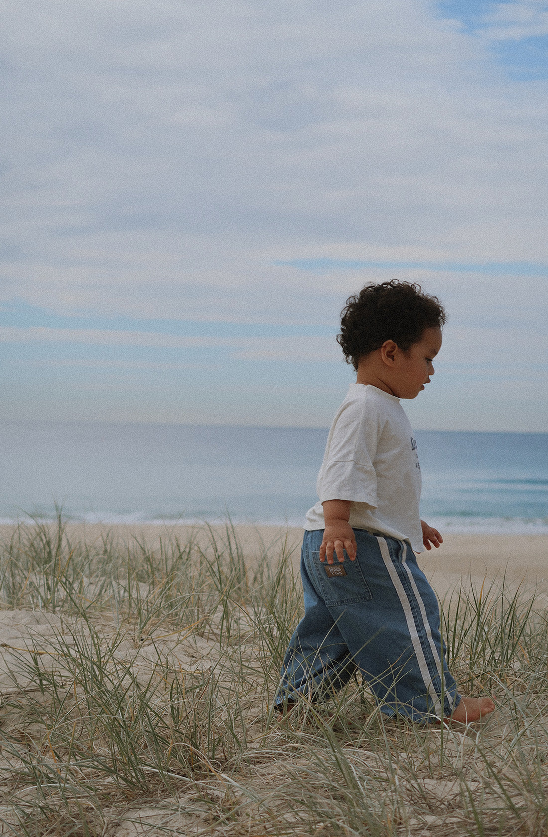 A young child with curly hair, wearing a white t-shirt and TWIN COLLECTIVE's Tricky Track Pant featuring a vintage-inspired design, walks barefoot through grass and sand dunes by the beach. The ocean and a partly cloudy sky are visible in the background.