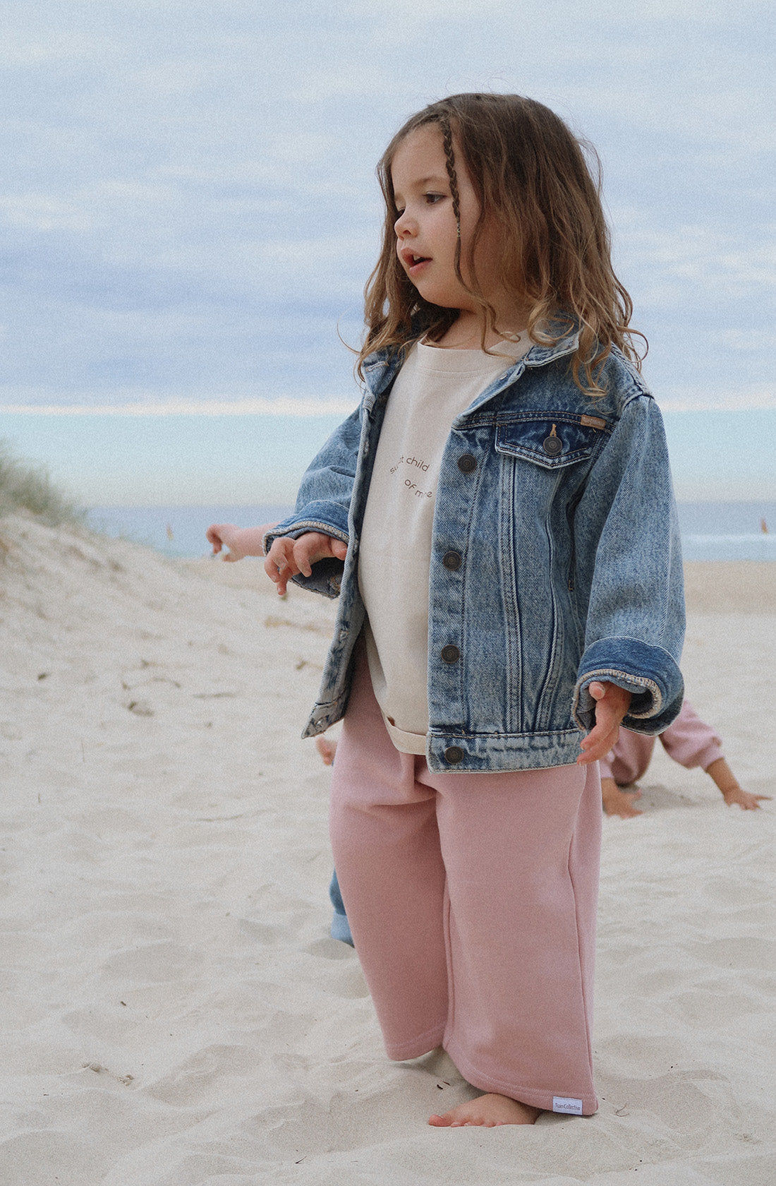 A young child with long hair stands on a sandy beach, sporting an oversized fit denim jacket over the TWIN COLLECTIVE Sweet Child Tee and pink pants. An overcast sky looms in the background, while the ocean stretches out behind them and another person can be seen partially, playing in the sand.