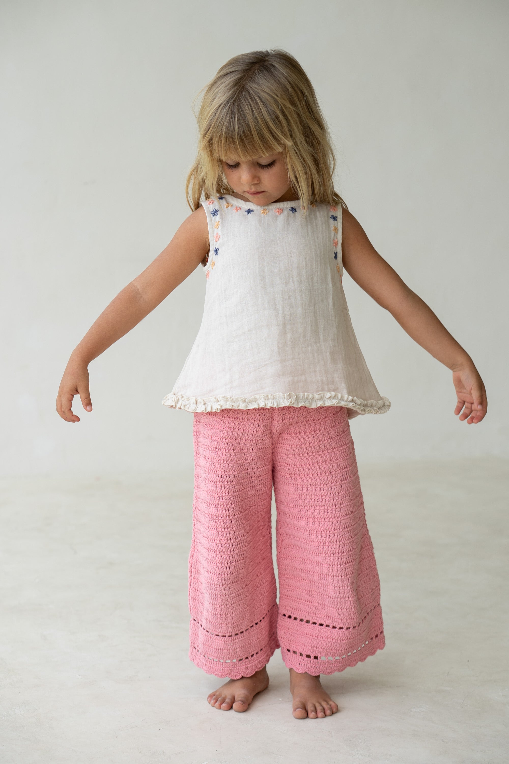 A young child with blonde hair stands barefoot on a light floor, wearing the ILLOURA THE LABEL Tilly Blouse Embroidered, a white sleeveless swing top adorned with colorful embroidery and floral trim around the neckline, paired with pink crochet pants. The airy cotton voile fabric complements the child's relaxed pose as they look down with arms slightly extended to the sides. The background is plain and light-colored.