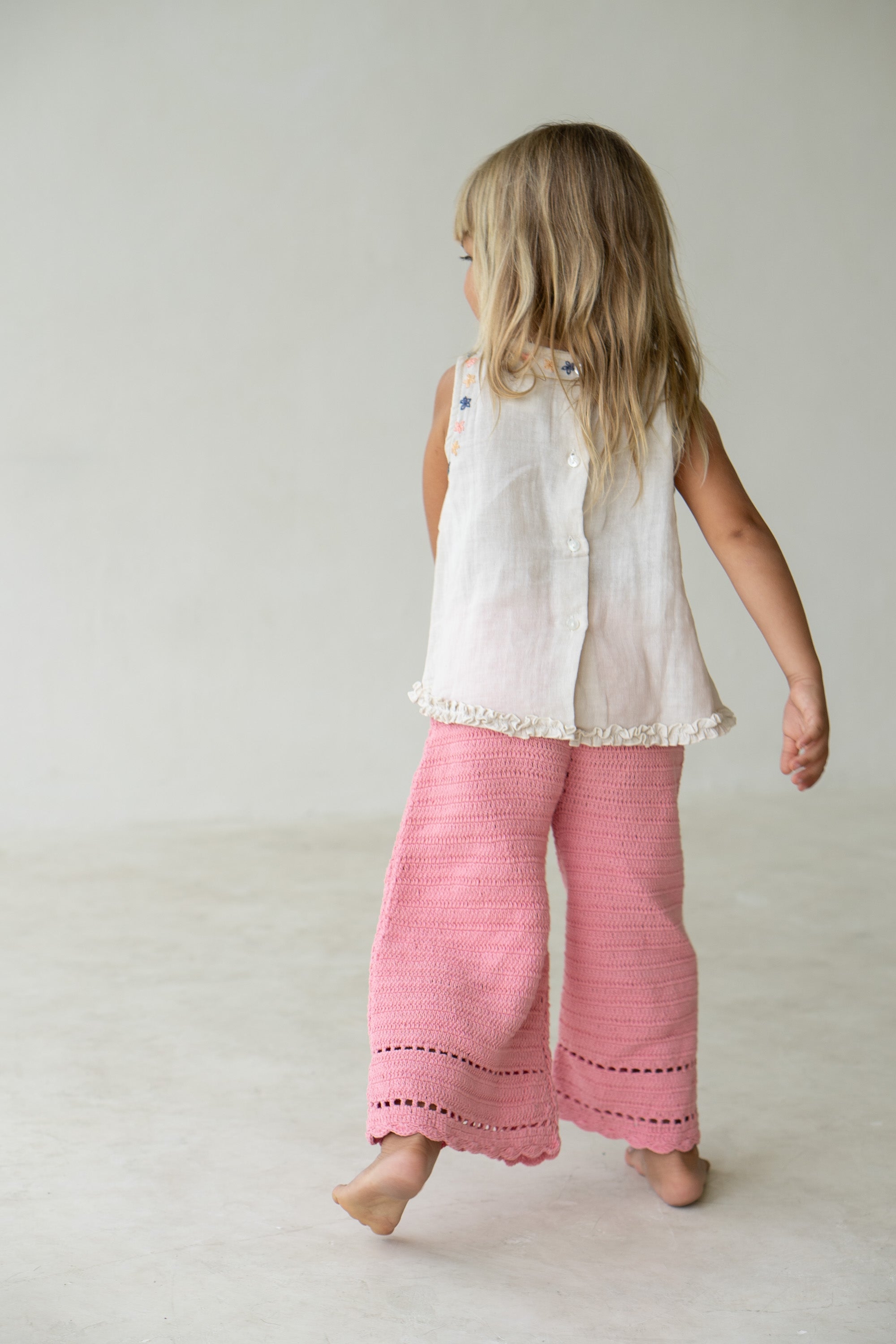 A young blonde girl, barefoot on a light-colored floor, stands facing away while appearing to be mid-step with one arm slightly extended behind her. She is wearing a white sleeveless top and the Crochet Pants Coral from ILLOURA THE LABEL, which have an elasticated waistband and a pink crocheted design.