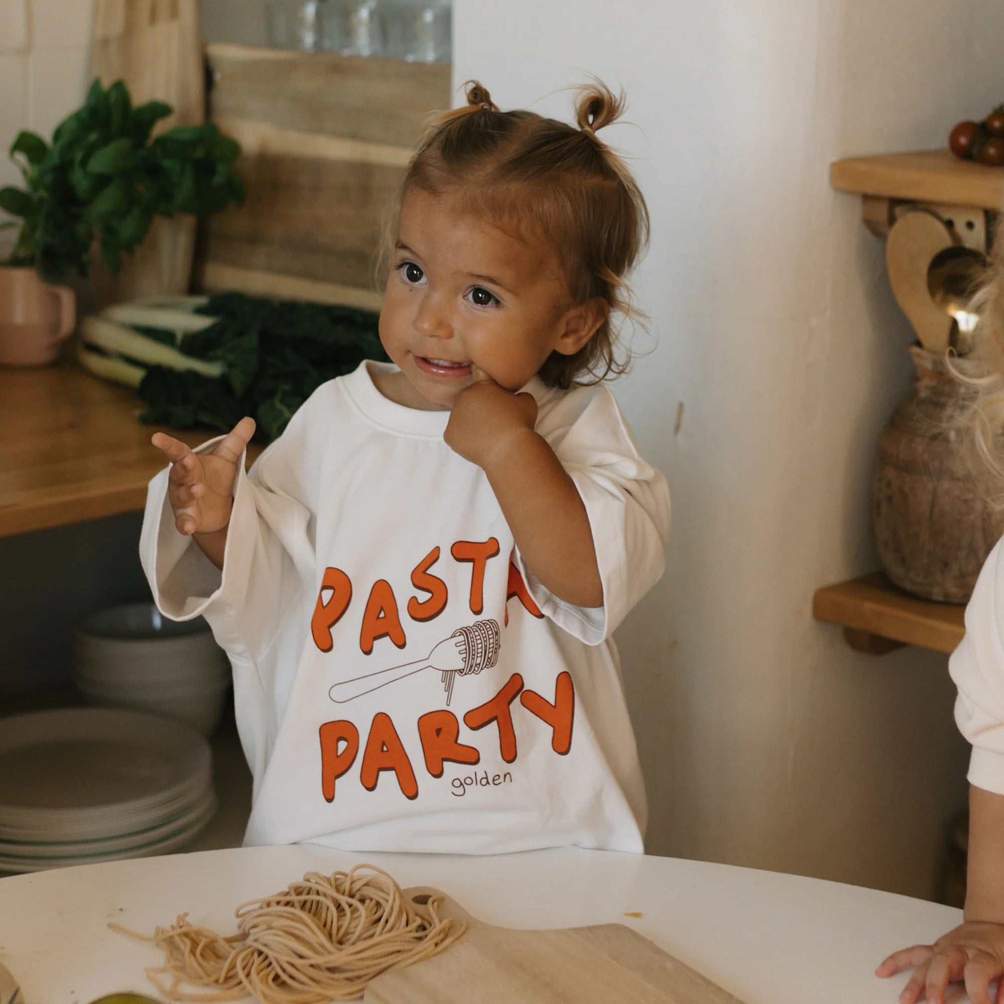 A delighted toddler with light brown hair in small buns stands in a kitchen wearing an oversized GOLDEN CHILDREN Pasta Party Mid Sleeve Tee Marshmallow, surrounded by uncooked pasta, a rolling pin, and lush greenery.