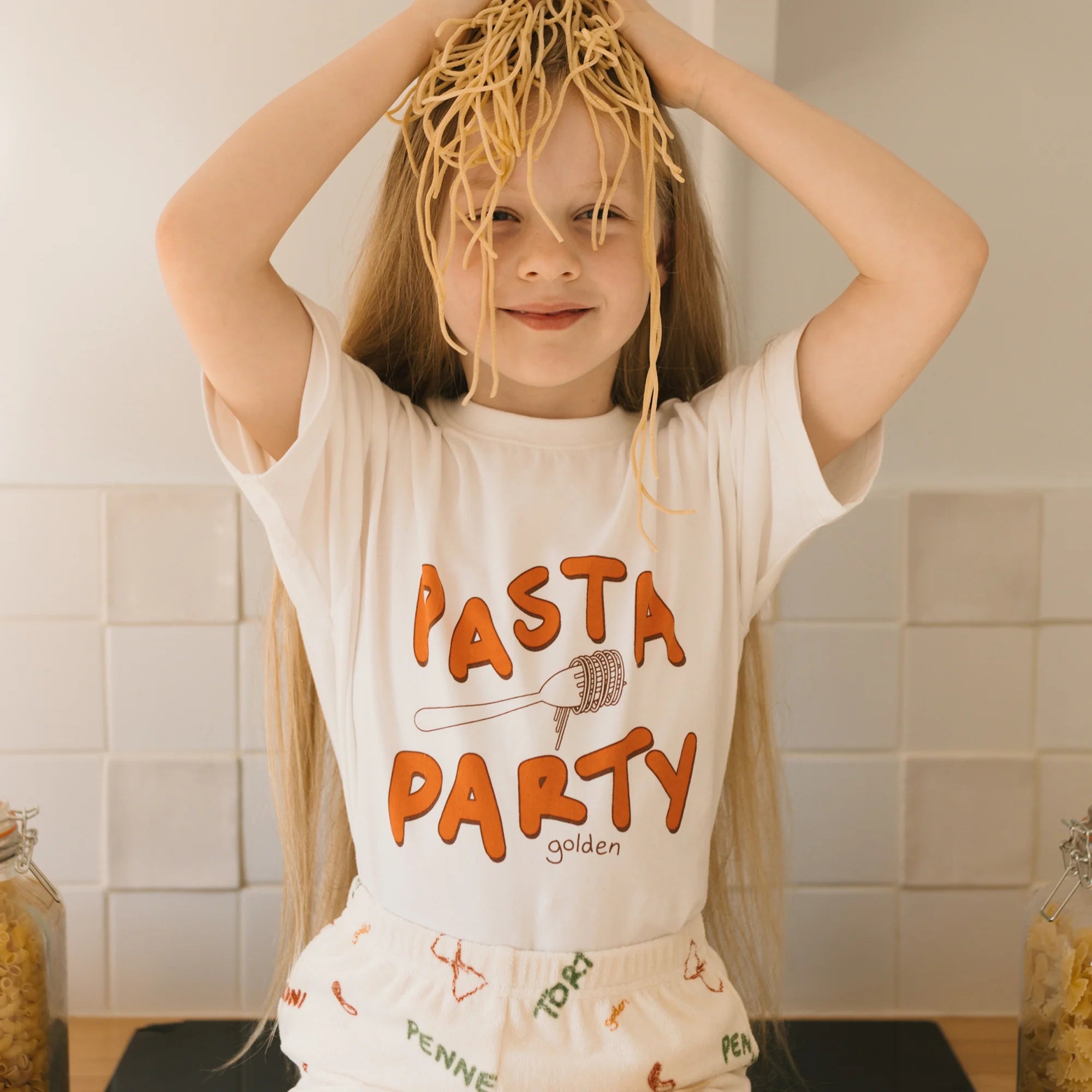 A child with long hair playfully wears a GOLDEN CHILDREN Pasta Party Mid Sleeve Tee Marshmallow, placing cooked spaghetti on their head. The oversized fit adds to the fun, with "Pasta Party" boldly featured, set against a kitchen backdrop filled with jars of pasta.