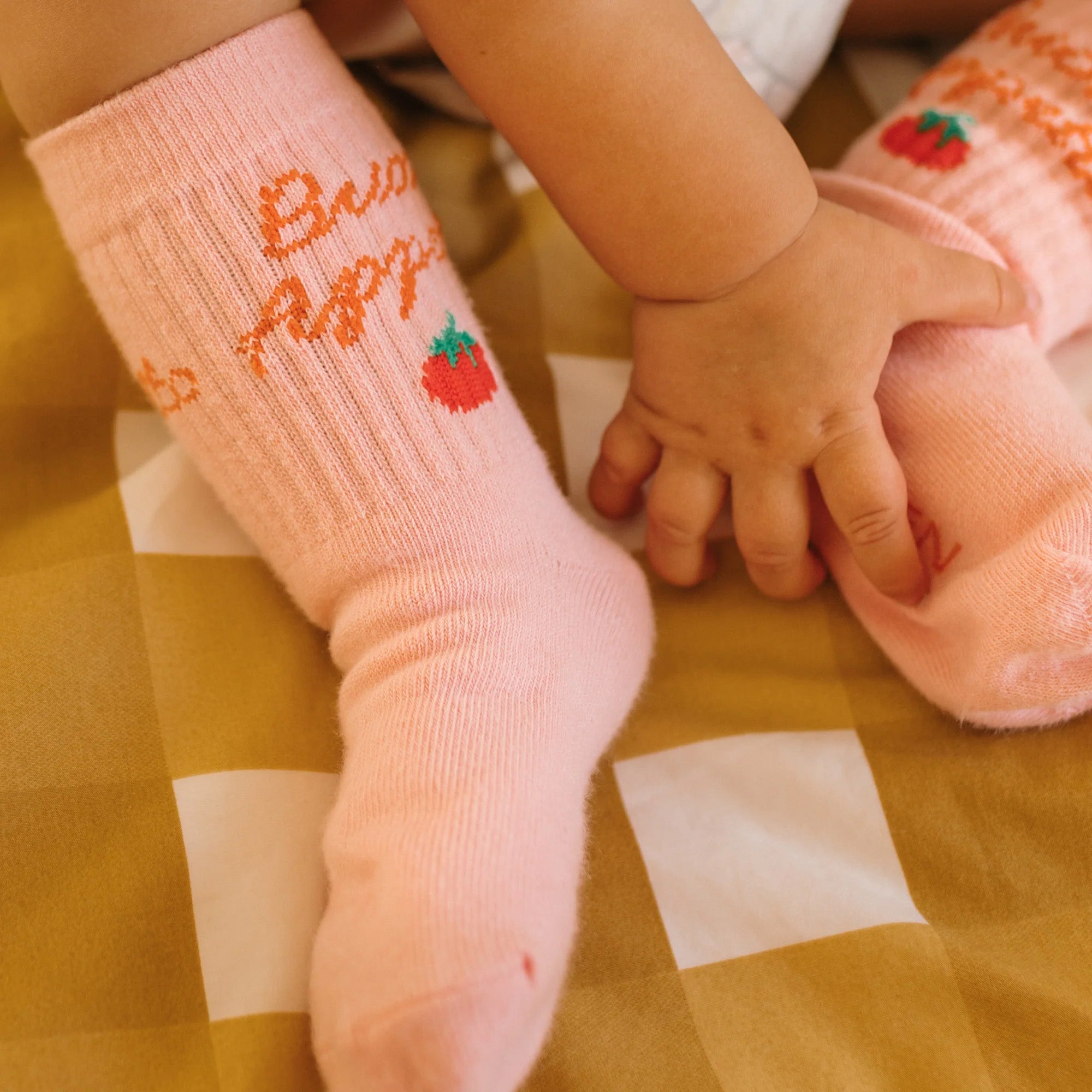 A baby wearing GOLDEN CHILDREN's Buon Appetito Socks Pink Parfait, featuring light pink ribbing with red apple designs and the text "Cute as an apple," sits on a checkered yellow and white blanket, hands touching one socked foot.