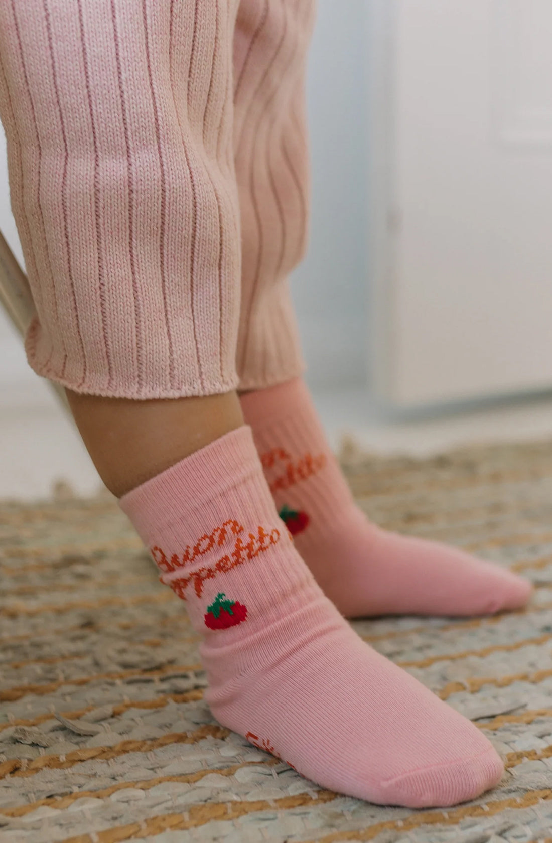 A person wearing pink ribbed pants and GOLDEN CHILDREN's Buon Appetito Socks Pink Parfait, adorned with small strawberry designs, stands on a woven rug. The setting appears to be indoors, with a light and airy background.