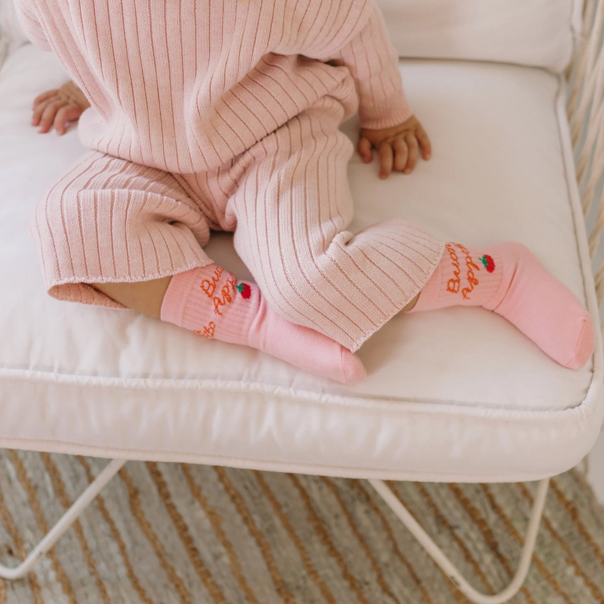 A baby dressed in a soft pink ribbed outfit sits on a cushioned chair, wearing GOLDEN CHILDREN's Buon Appetito Socks Pink Parfait adorned with playful embroidery. The chair is placed on a beige and white striped rug, creating an overall cozy and comfortable setting.