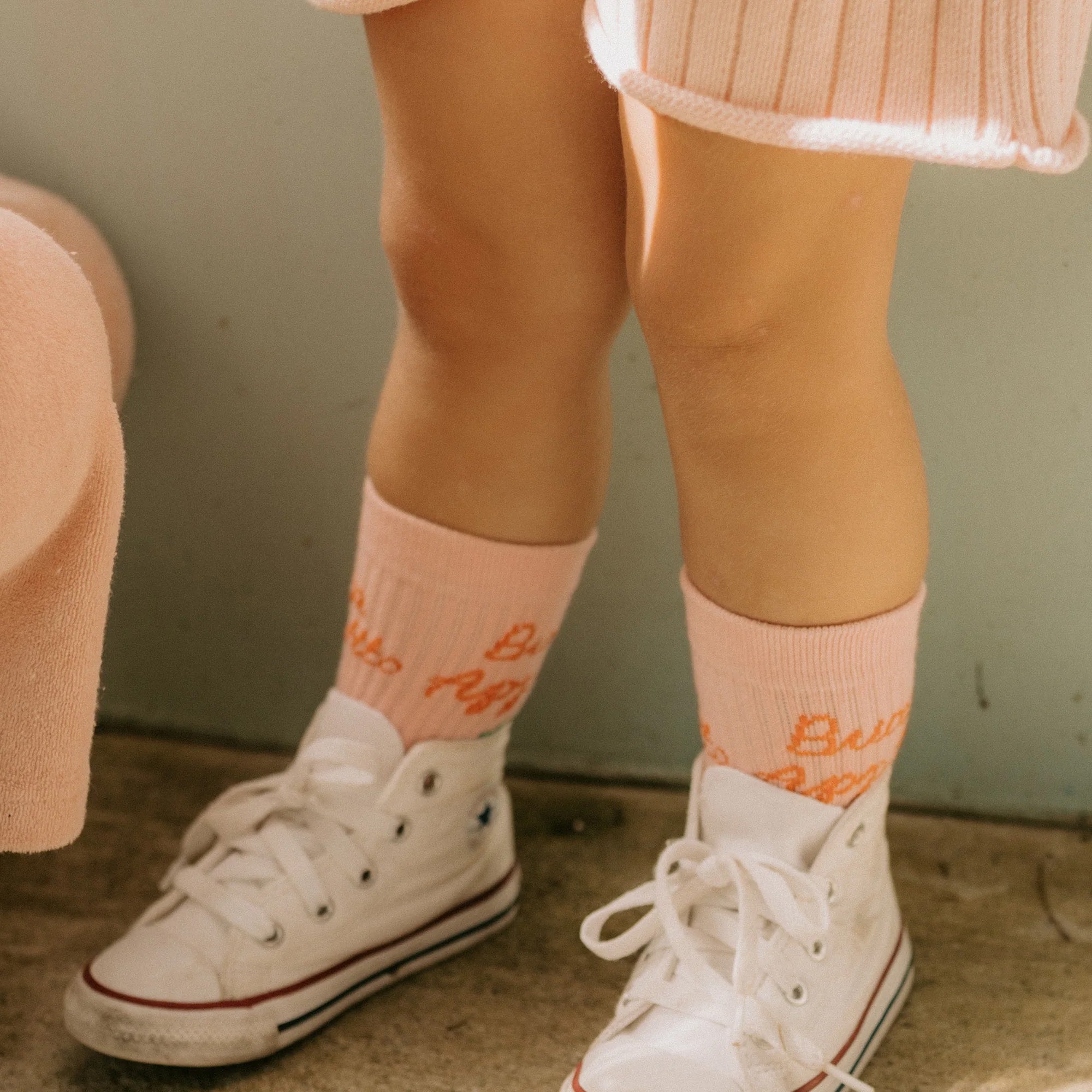 A child wearing light pink Buon Appetito Socks Pink Parfait by GOLDEN CHILDREN, adorned with "Baby" lettering, pairs them with white high-top sneakers that have reinforced toe caps. The child is dressed in light-colored clothing, with their legs visible in the frame, standing on a concrete floor near a light green wall.