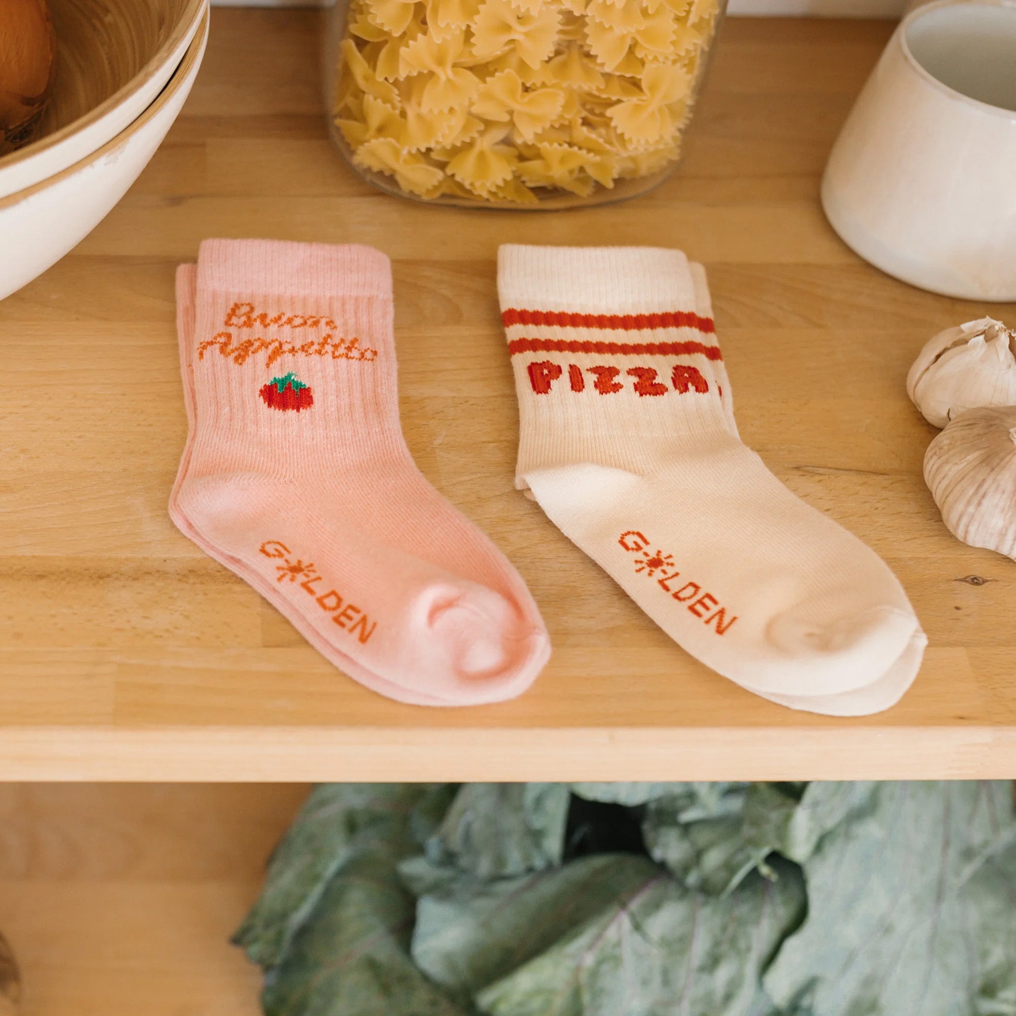 Two pairs of Buon Appetito Socks Pink Parfait are displayed on a wooden surface among pasta and vegetables. The left sock is pink with the phrase "Buon Appetito" and a red tomato design, while the right sock is beige with the word "PIZZA" in red letters. Both feature reinforced toe caps and have the "GOLDEN CHILDREN" brand name on the toes.