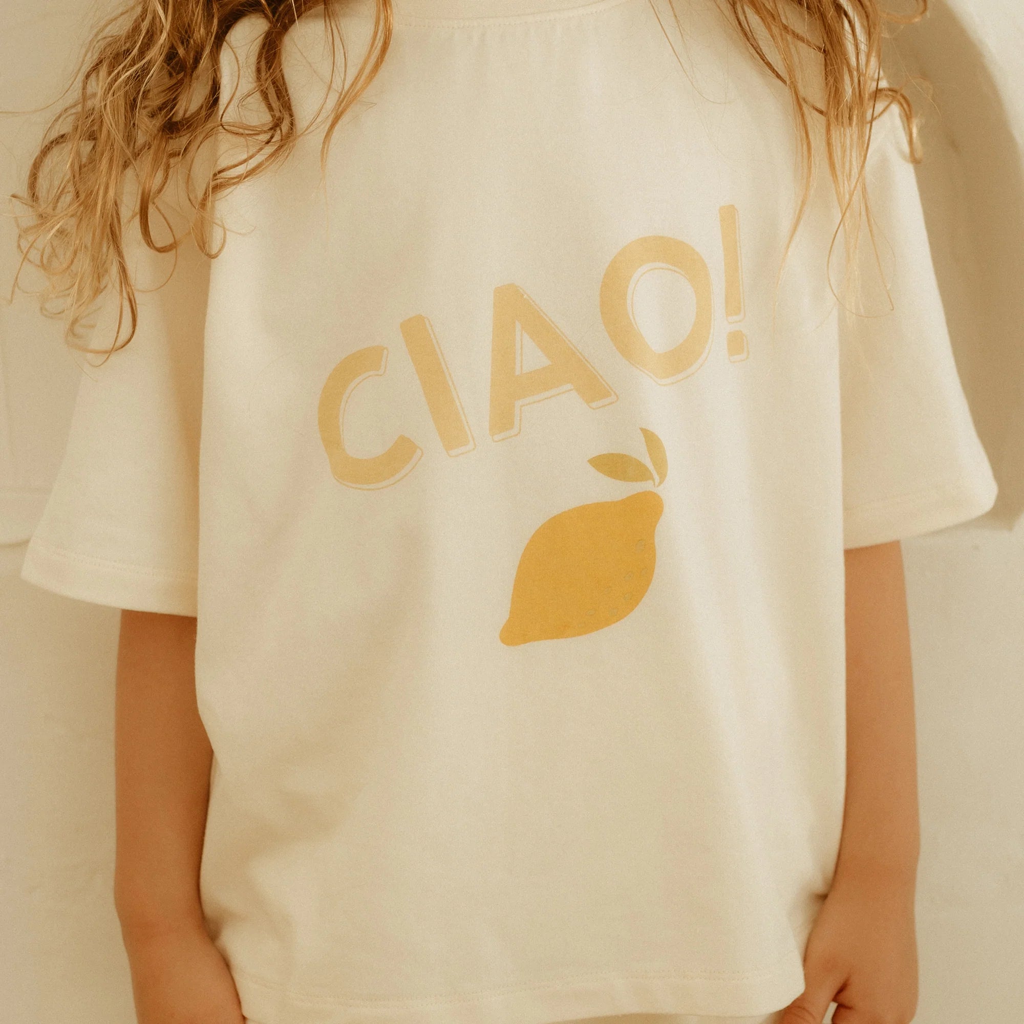 A child wearing a relaxed-fit Ciao Mid Sleeve Tee Buttercream by GOLDEN CHILDREN, crafted from soft cotton jersey, features the bright word "CIAO!" with a cheerful yellow lemon graphic. The child's long, wavy hair gently cascades down their back.