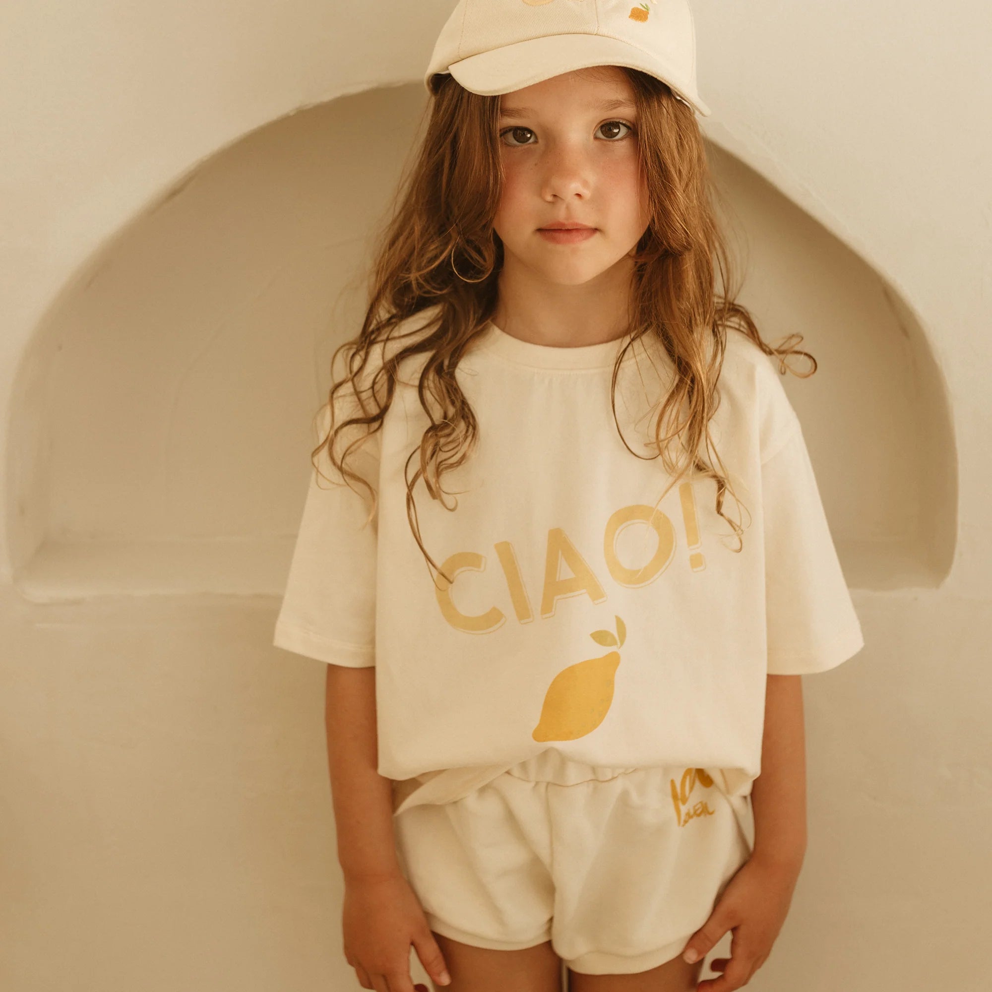 A young girl poses against a white arched backdrop, wearing the GOLDEN CHILDREN's Ciao Mid Sleeve Tee in Buttercream with a lemon graphic. Her relaxed-fit cotton jersey tee is paired with a matching cap, and her long hair flows freely as she confidently gazes at the camera.