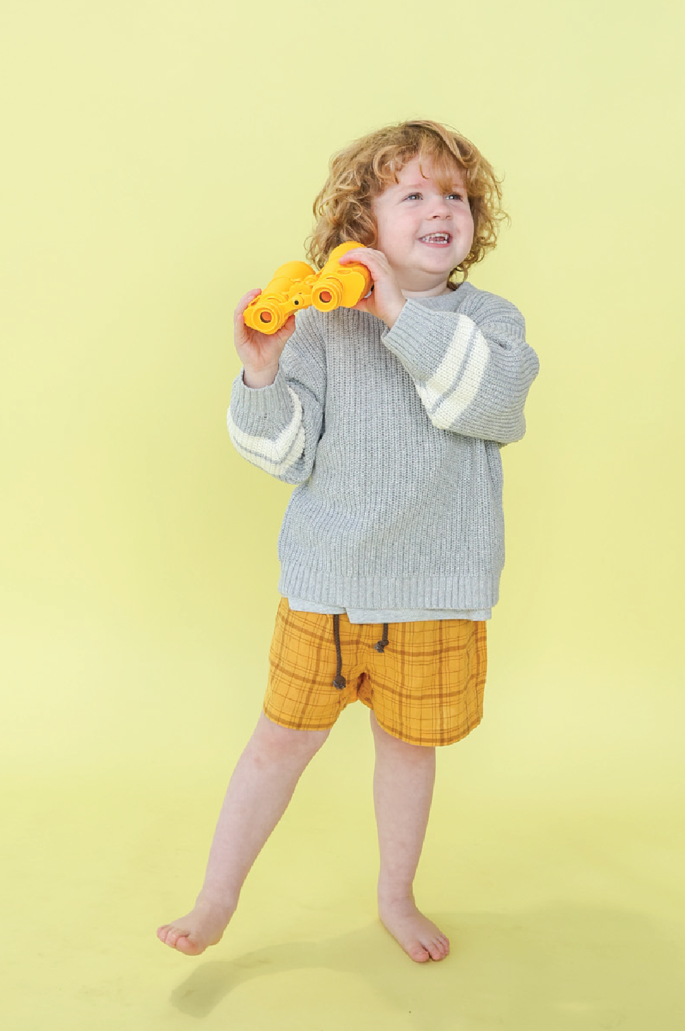 A young child with curly hair stands barefoot on a yellow background, smiling and holding yellow toy binoculars. They wear a gray sweater with white stripes and GROWN - Plaid Shorts crafted from lightweight woven check fabric.