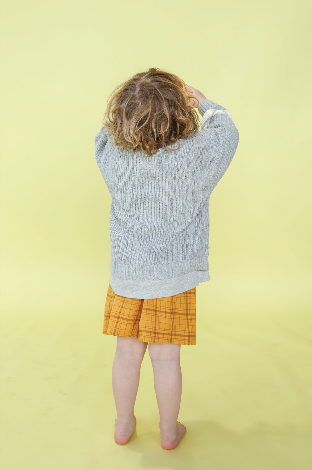 A child with curly hair, barefoot on a yellow background and facing away from the camera, wears GROWN's Plaid Shorts and a grey sweater. The shorts feature lightweight woven check fabric and a drawstring waistband.