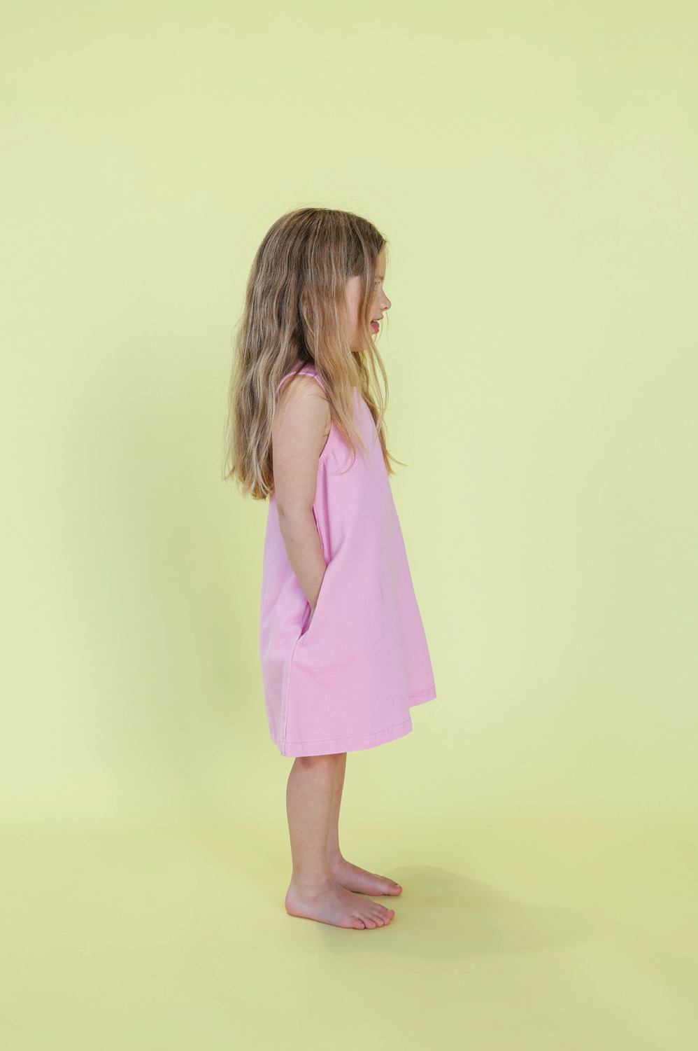 A young girl with long light brown hair stands barefoot in profile, wearing the GROWN Organic Shift Denim Dress Candy, hands in her pockets, against a pale yellow background, looking to the right.