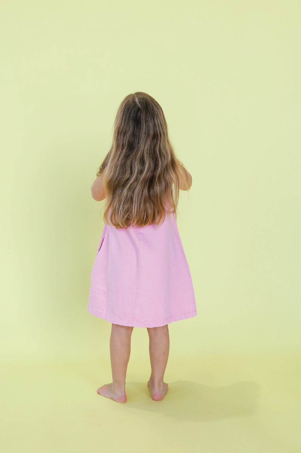A young girl with long brown hair wears the GROWN Organic Shift Denim Dress in Candy, standing barefoot and facing away from the camera against a plain yellow background.
