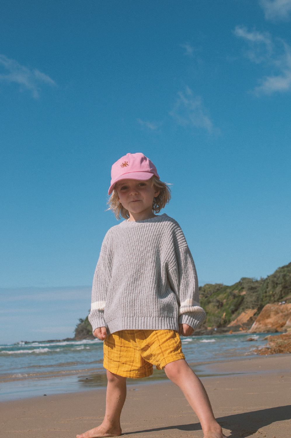 A young child in a pink cap, gray sweater, and GROWN Plaid Shorts stands barefoot on a sandy beach, with the ocean and greenery behind them under a clear blue sky.