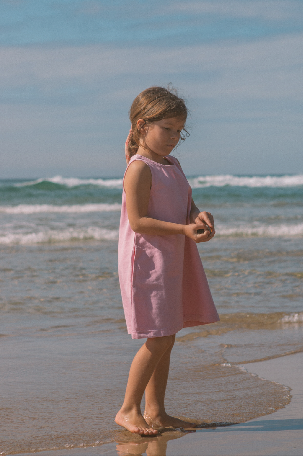 A young girl wearing the GROWN Organic Shift Denim Dress Candy stands barefoot on wet sand by the ocean, looking down at something in her hands as gentle waves and a blue sky form the backdrop.