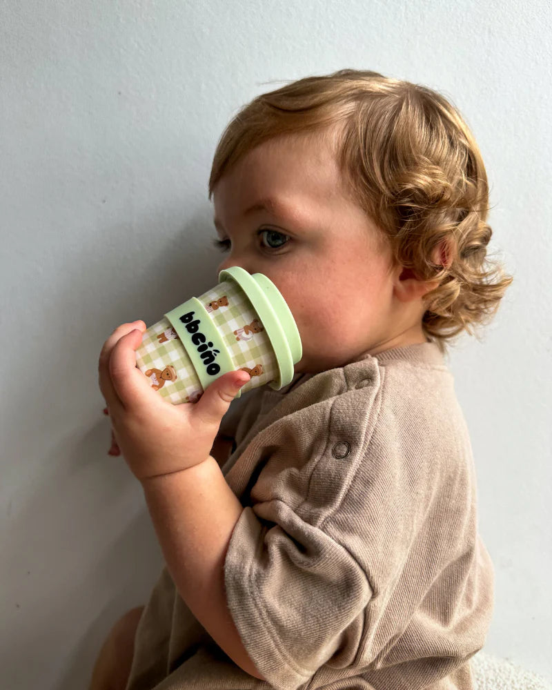 A young child with curly hair sips from a BBCINO BBcino ~ Reusable Babycino Cup 120ml Cub, featuring bear designs and "bebeino," while wearing a beige shirt against a light gray background, highlighting the charm of sustainably farmed bamboo cups.