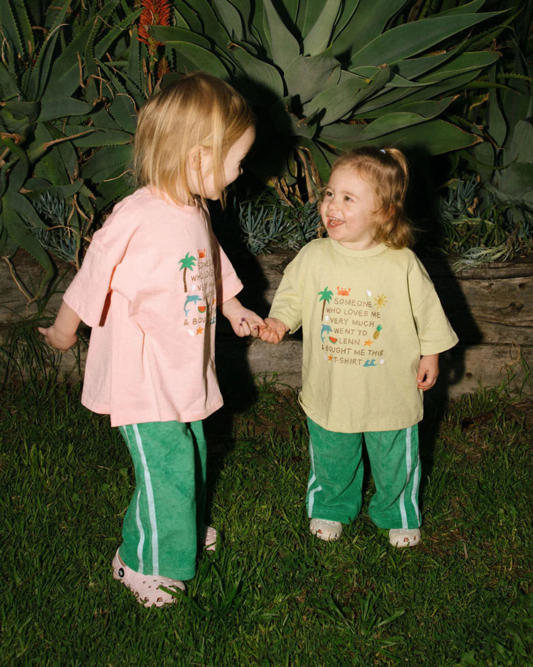 Two young children smile and hold hands on grass, wearing the LENN LABEL Lenn Tee Blush. Their relaxed fit cotton tees stand out as they play among lush leafy plants, creating a warm and playful scene.