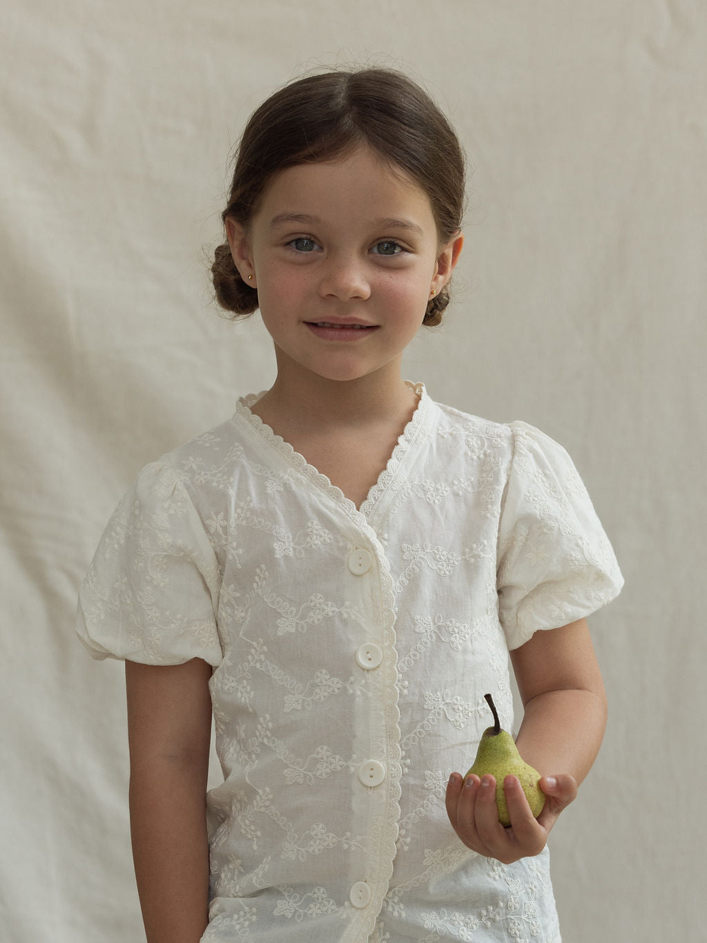 A young girl with dark hair tied in pigtails stands against a light background, wearing the Hinterland Blouse by VALENCIA BYRON BAY, adorned with floral embroidered details. She smiles gently while holding a small pear in her right hand, exuding a charming innocence.