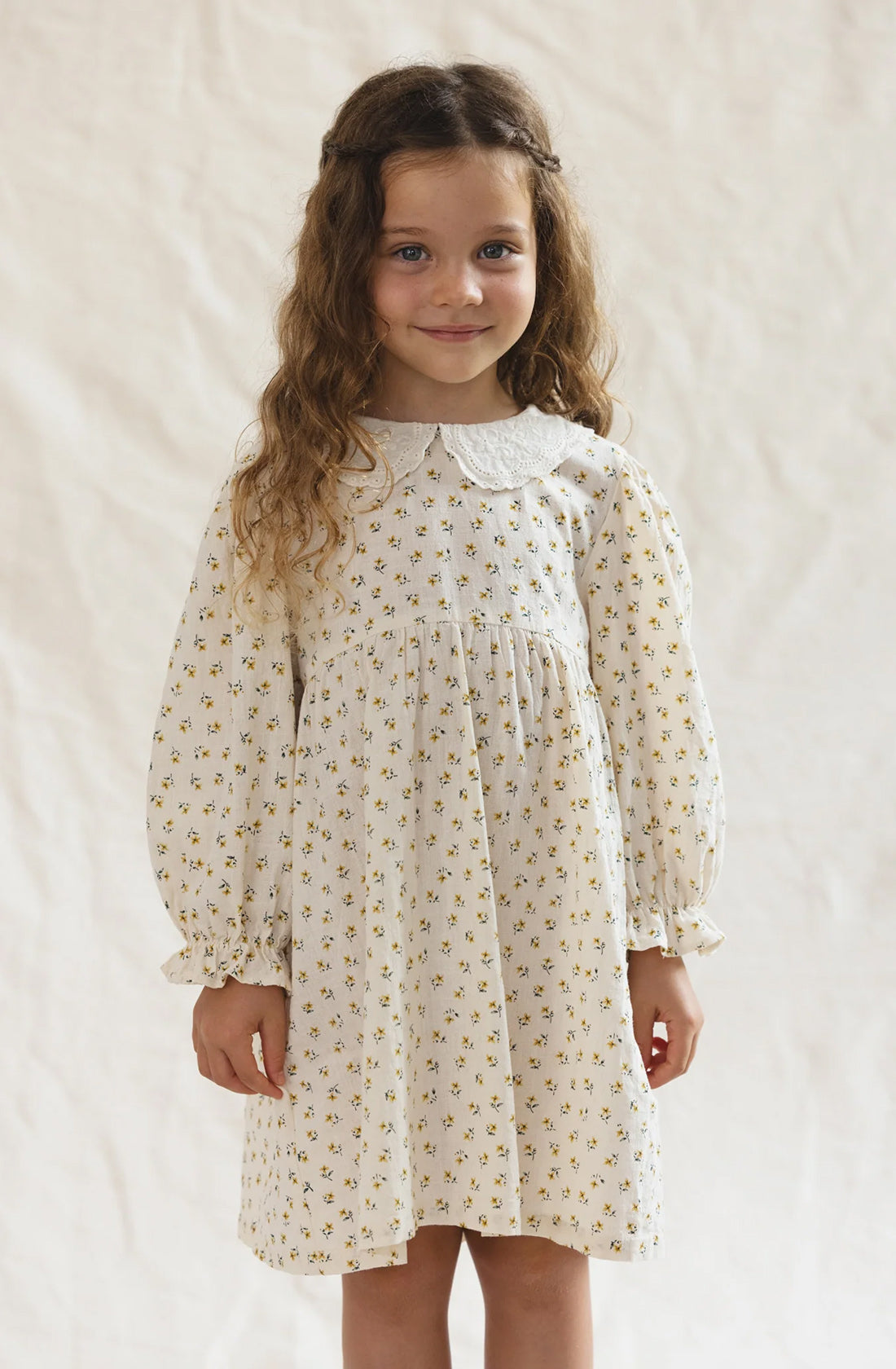 A young girl stands against a light background, smiling softly. She has long, wavy brown hair and is wearing The Farmer's Daughter's Dress by VALENCIA BYRON BAY, a cream-colored outfit adorned with a floral pattern, Peter Pan collar, and gathered cuffs on the long sleeves. The classic style is complemented by the breathable mid-weight fabric.