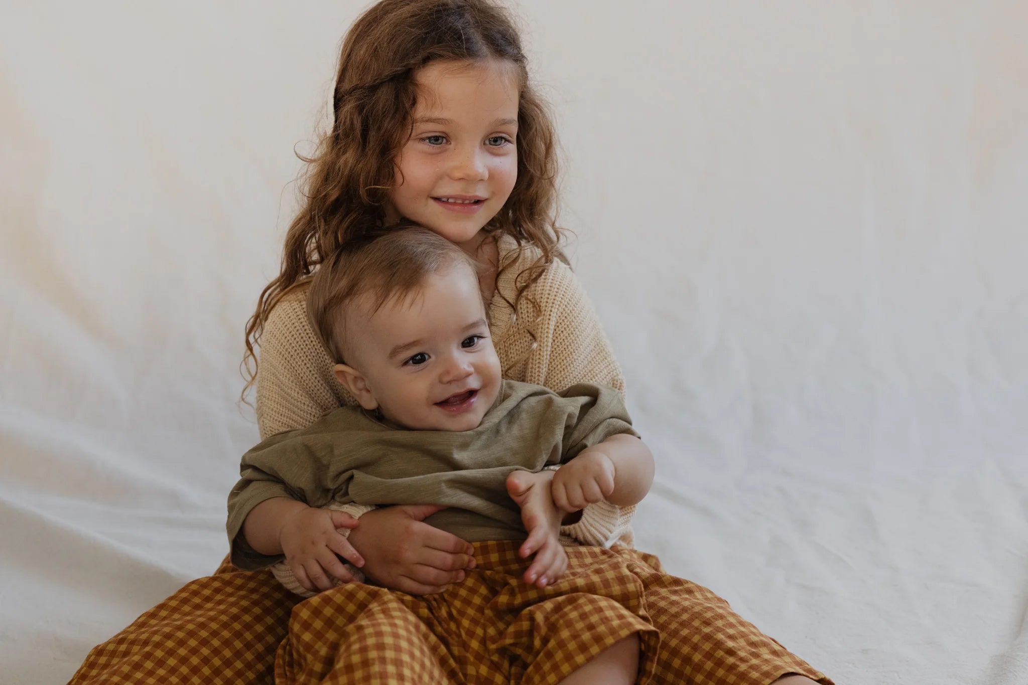 A young girl with wavy brown hair and a warm smile is sitting on a neutral-colored backdrop, holding a happy baby in her lap. Both children are dressed in beige and brown clothing, including the versatile Rio Pant Honeycomb from VALENCIA BYRON BAY, made from 100% sand washed cotton. The baby's clapping hands add to the joyous moment as the girl supports him lovingly.