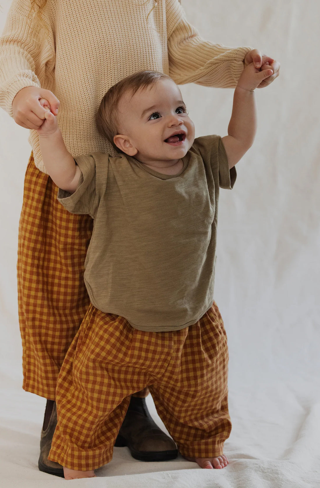 Two children are standing together, holding hands. The younger child, barefoot and smiling, is wearing a green shirt and mustard Rio Pant Honeycomb from Valencia Byron Bay made from 100% sand-washed cotton. The older child, whose face is not visible, sports a beige sweater and similar checkered pants in the same unisex design.