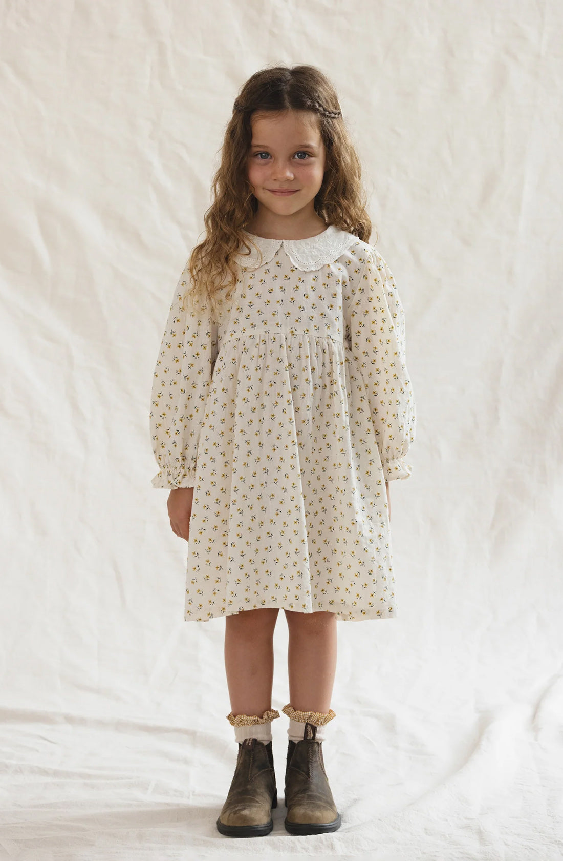 A young girl with long, wavy hair stands against a plain, light-colored background. She wears the classic style of The Farmer's Daughter's Dress from VALENCIA BYRON BAY, featuring white fabric adorned with a small floral pattern and a Peter Pan collar. Complementing her look are brown ankle boots and ruffled socks.