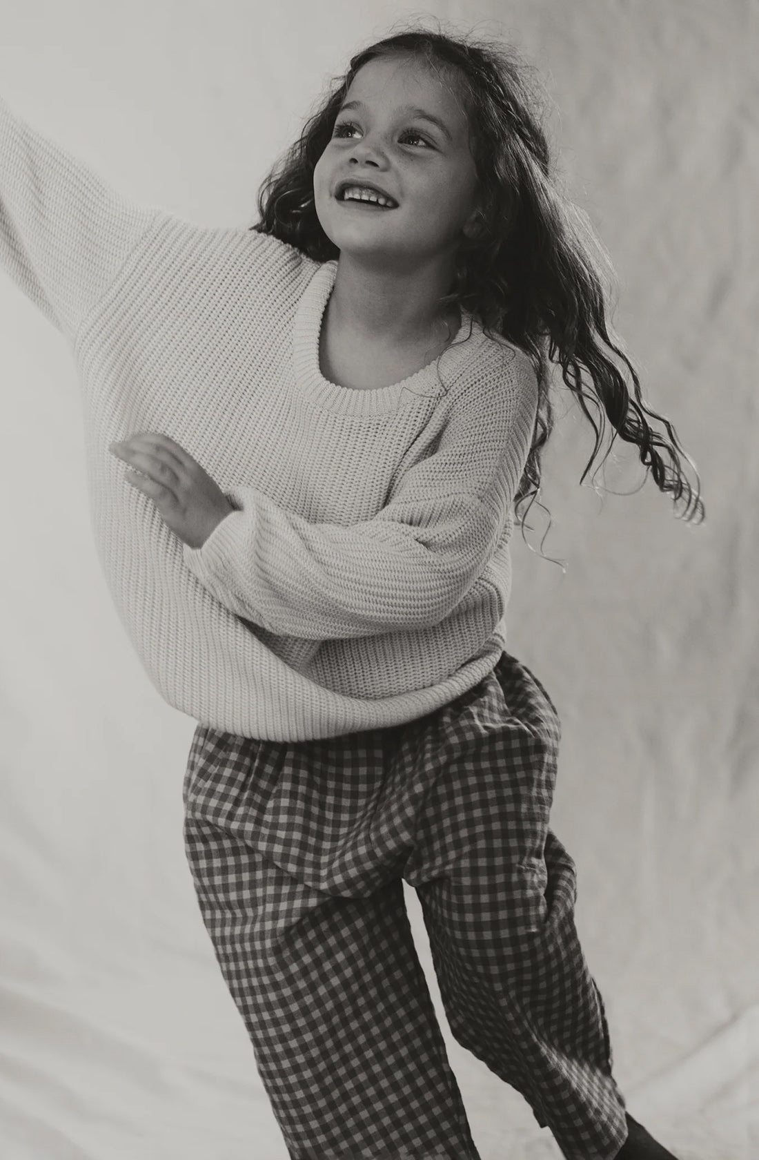 A young girl with curly hair, wearing a cozy knit sweater and the Rio Pant Honeycomb by VALENCIA BYRON BAY made from 100% sand-washed cotton, smiles and reaches out with one arm while her other arm is bent. She appears to be dancing or playfully posing against a simple, light-colored background. The image is in black and white.