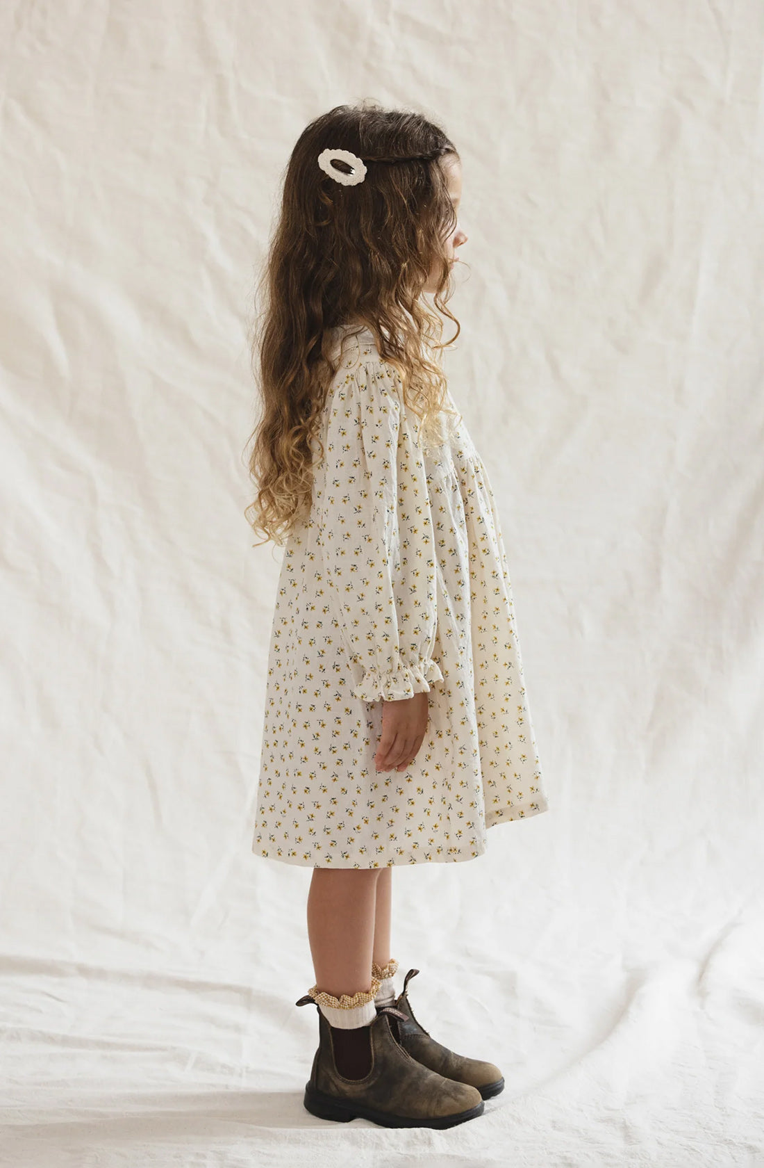 A young girl with long, wavy hair stands in profile against a plain background. She wears The Farmer's Daughter's Dress by VALENCIA BYRON BAY, crafted from breathable mid-weight fabric and adorned with a small floral pattern. Her outfit is completed with ankle socks and brown boots, and a white hair clip is visible in her hair.