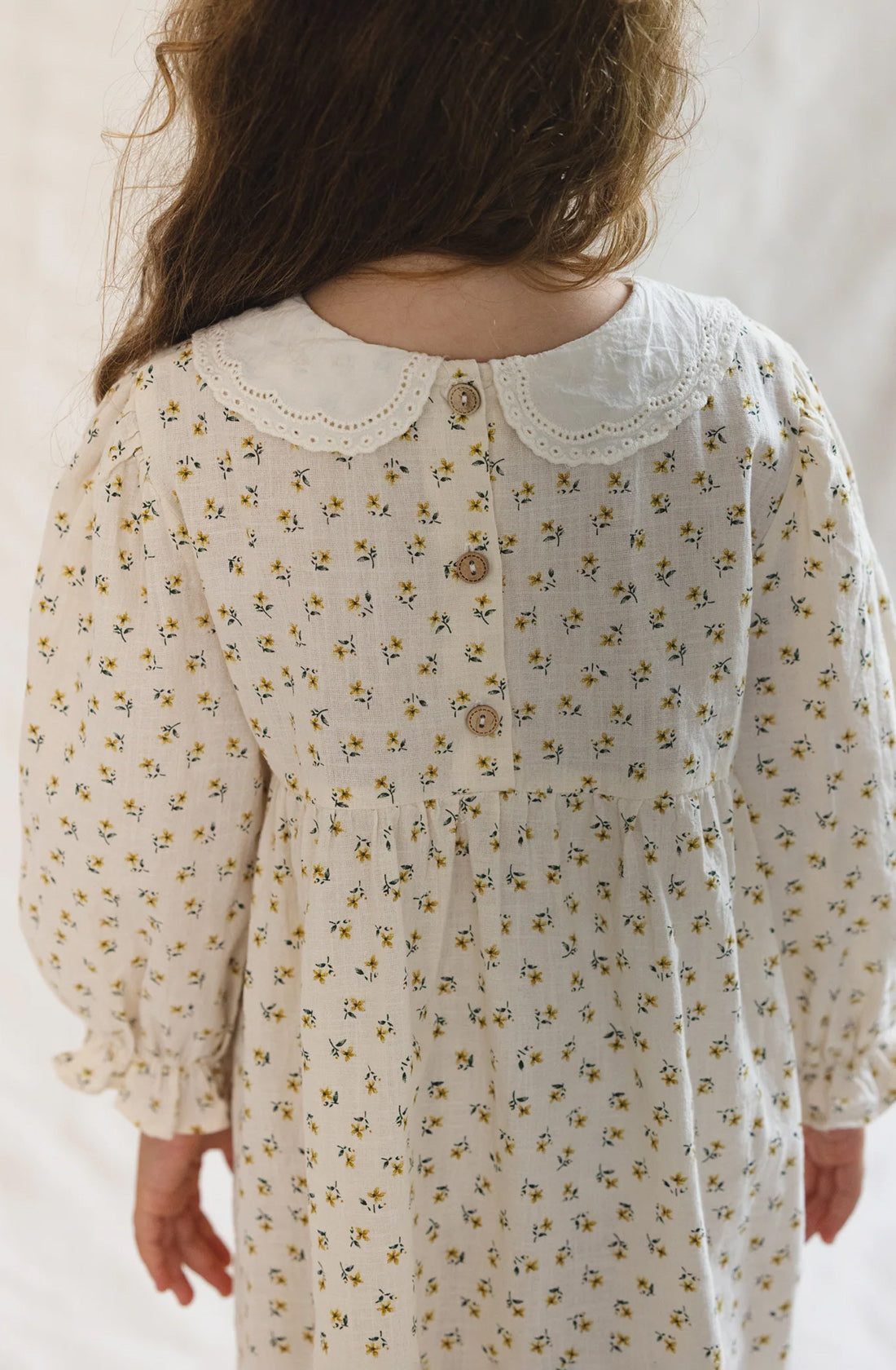 A young girl with long, wavy hair stands with her back to the camera, wearing The Farmer's Daughter's Dress by VALENCIA BYRON BAY—a cream-colored piece adorned with small yellow floral patterns. The classic style features a scalloped white lace collar and buttons down the back, crafted from breathable mid-weight fabric.