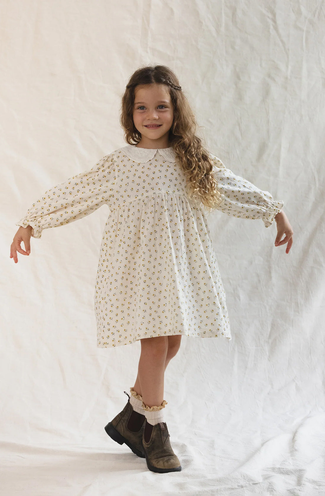 A young girl with long curly hair stands against a light fabric backdrop. She wears The Farmer's Daughter's Dress by VALENCIA BYRON BAY, a white long-sleeve piece with a floral pattern made from breathable mid-weight fabric, and brown ankle boots. She smiles and extends her arms slightly to the sides, appearing cheerful and playful.
