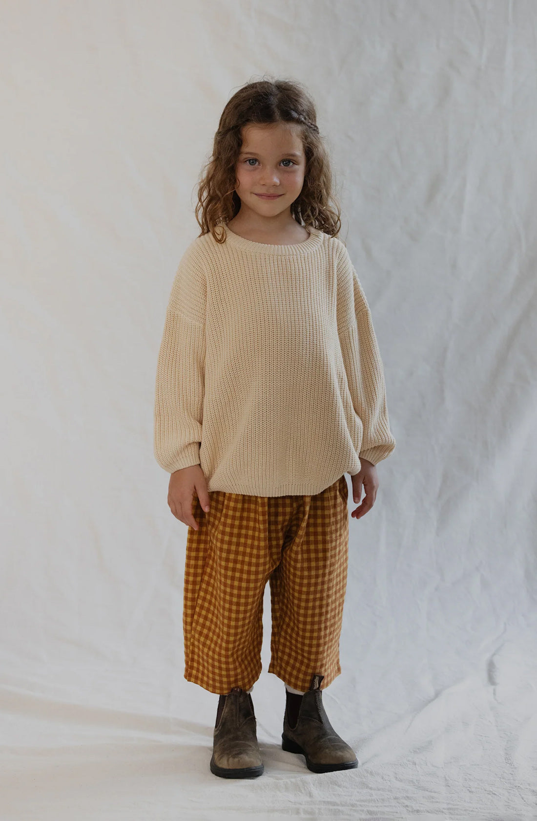 A young child with curly hair stands against a neutral backdrop. They are wearing an oversized beige sweater paired with Valencia Byron Bay's Rio Pant Honeycomb in a checkered mustard-yellow and brown design, complemented by brown boots. The unisex design ensures comfort, as the child's smile softens their expression, hands resting comfortably by their sides.