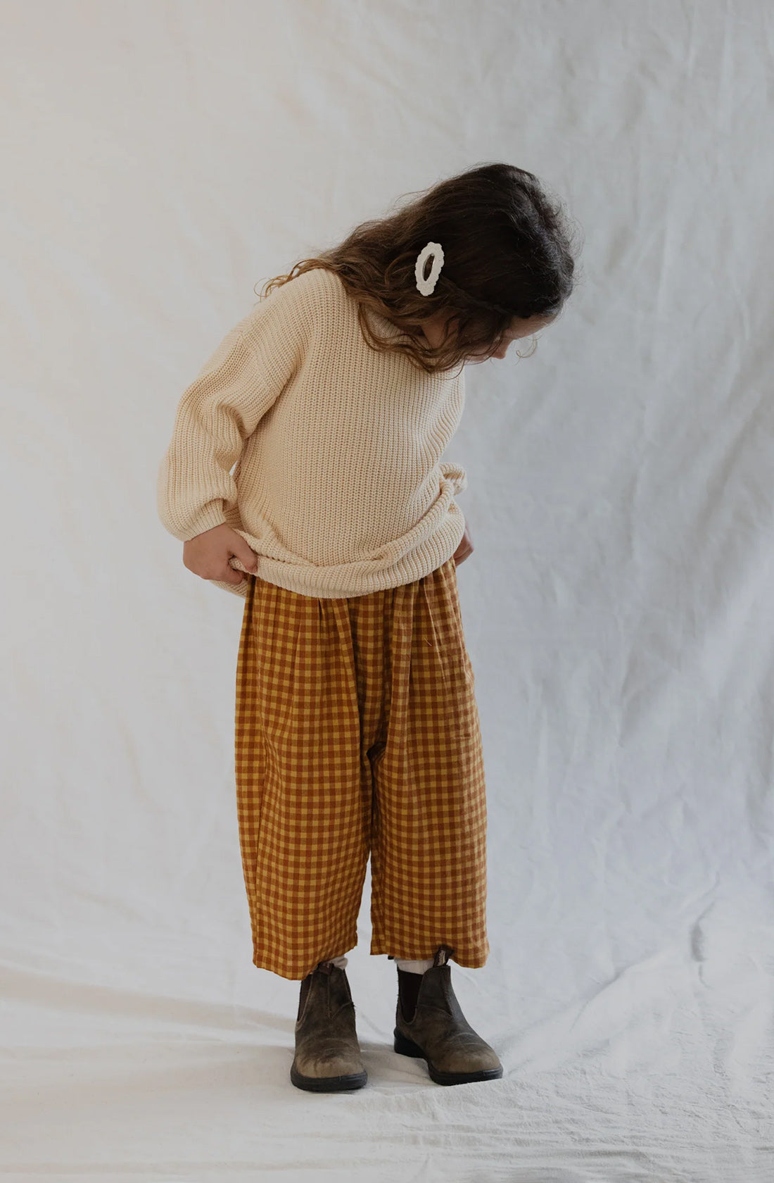 A young child with long hair stands against a light background, looking down. They are wearing a light cream sweater, Rio Pant Honeycomb in a mustard and brown checkered pattern made from 100% sand washed cotton by VALENCIA BYRON BAY, brown boots, and a white hairclip.